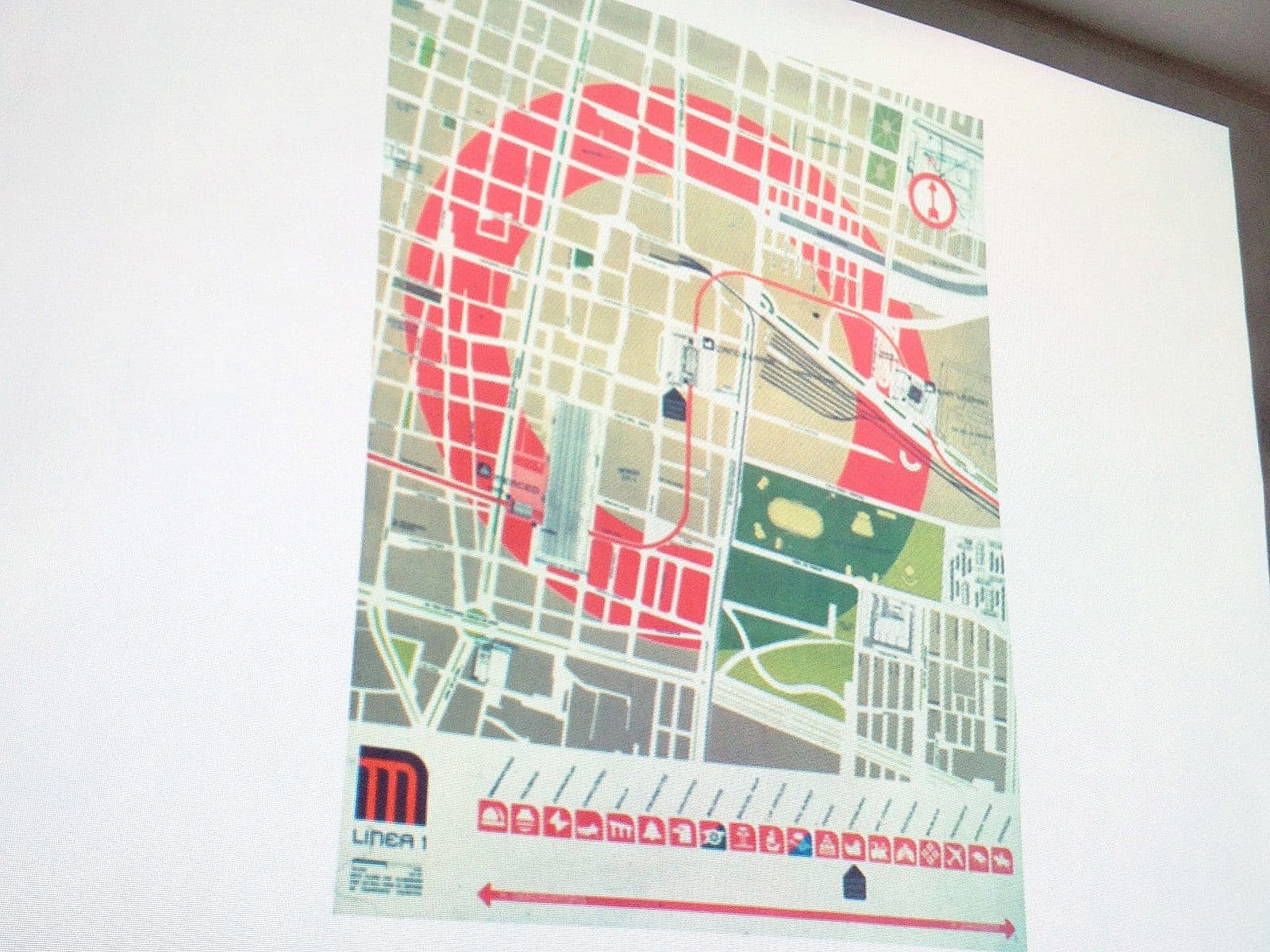 A projected image displaying a map of a city with a prominently marked red arc representing Line 1 of a metro system. Various streets, blocks, and green areas are visible, with metro symbols and station names along the red line at the bottom of the map.