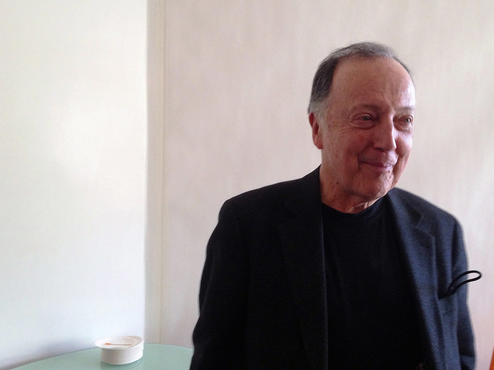 An older man with short, dark hair, dressed in a black suit jacket and black shirt, stands in a well-lit room. He is smiling and looking slightly to his right. A white table with a small white bowl and some items on it is seen in the background.