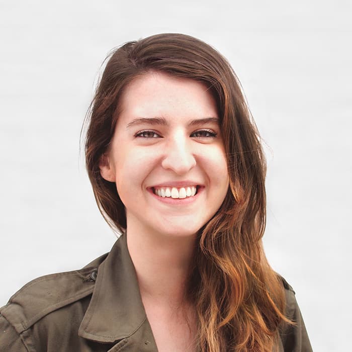 A person with long, wavy brown hair, wearing an olive green jacket, smiles warmly at the camera. The background is a plain, light gray color.