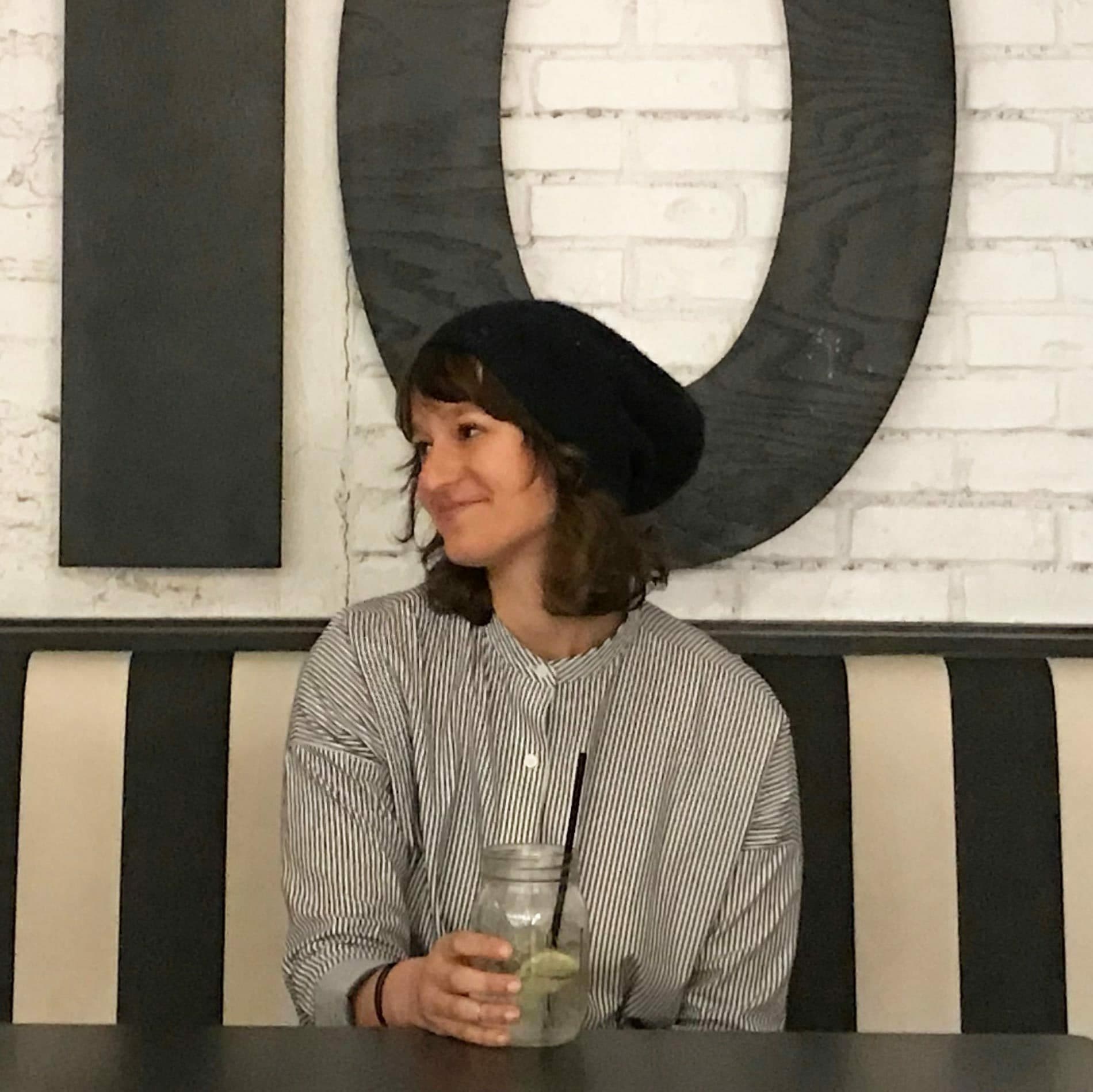 A smiling woman wearing a black beanie and a striped shirt is sitting on a black and white striped bench with a mason jar filled with a drink in front of her. She is looking to her right and there is a large decorative letter 'O' behind her on a white brick wall.