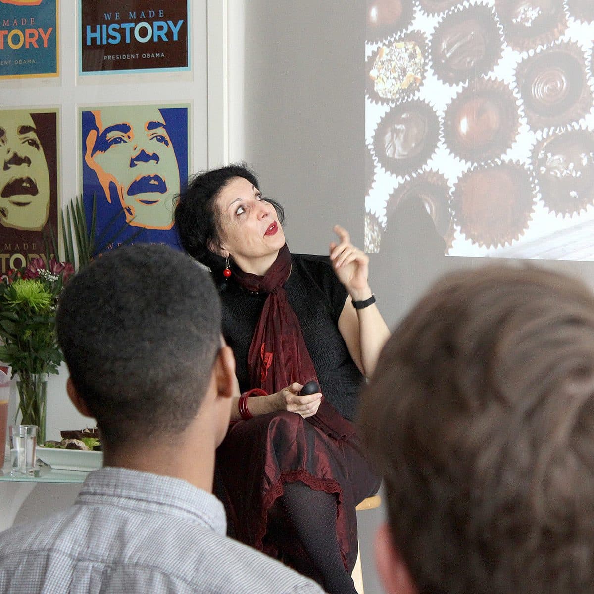 A woman dressed in a dark outfit gestures and speaks to a seated audience in a classroom. Behind her are posters featuring Barack Obama and a projected image of chocolates. To the side is a table with a pitcher of drink and flowers.