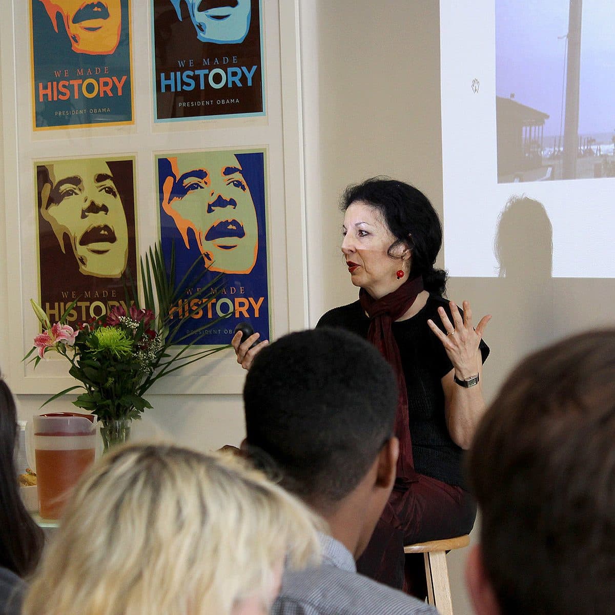 A woman sits on a stool, gesturing with her hands as she speaks to an audience. Behind her are posters of Barack Obama with the words "We Made History" and a projector screen displaying an image. A vase with flowers and a drink are on a table next to her.