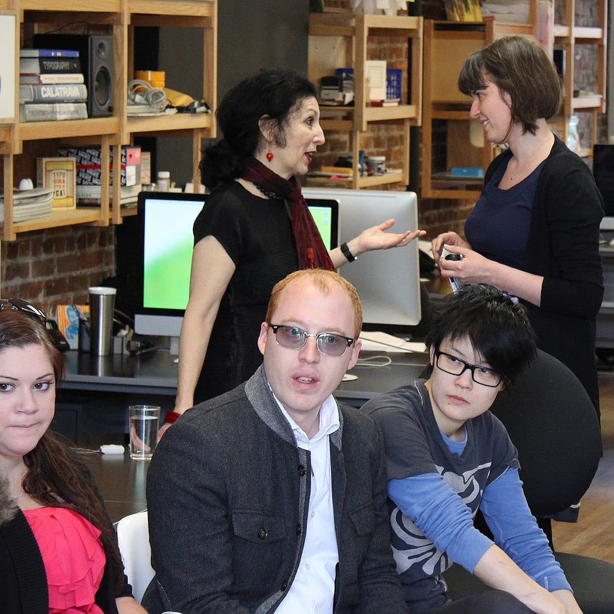 A group of people are inside an office space. In the foreground, three individuals are seated and facing the camera, while two others are standing behind them and chatting. The room has various computer equipment and shelves with items on the wall.