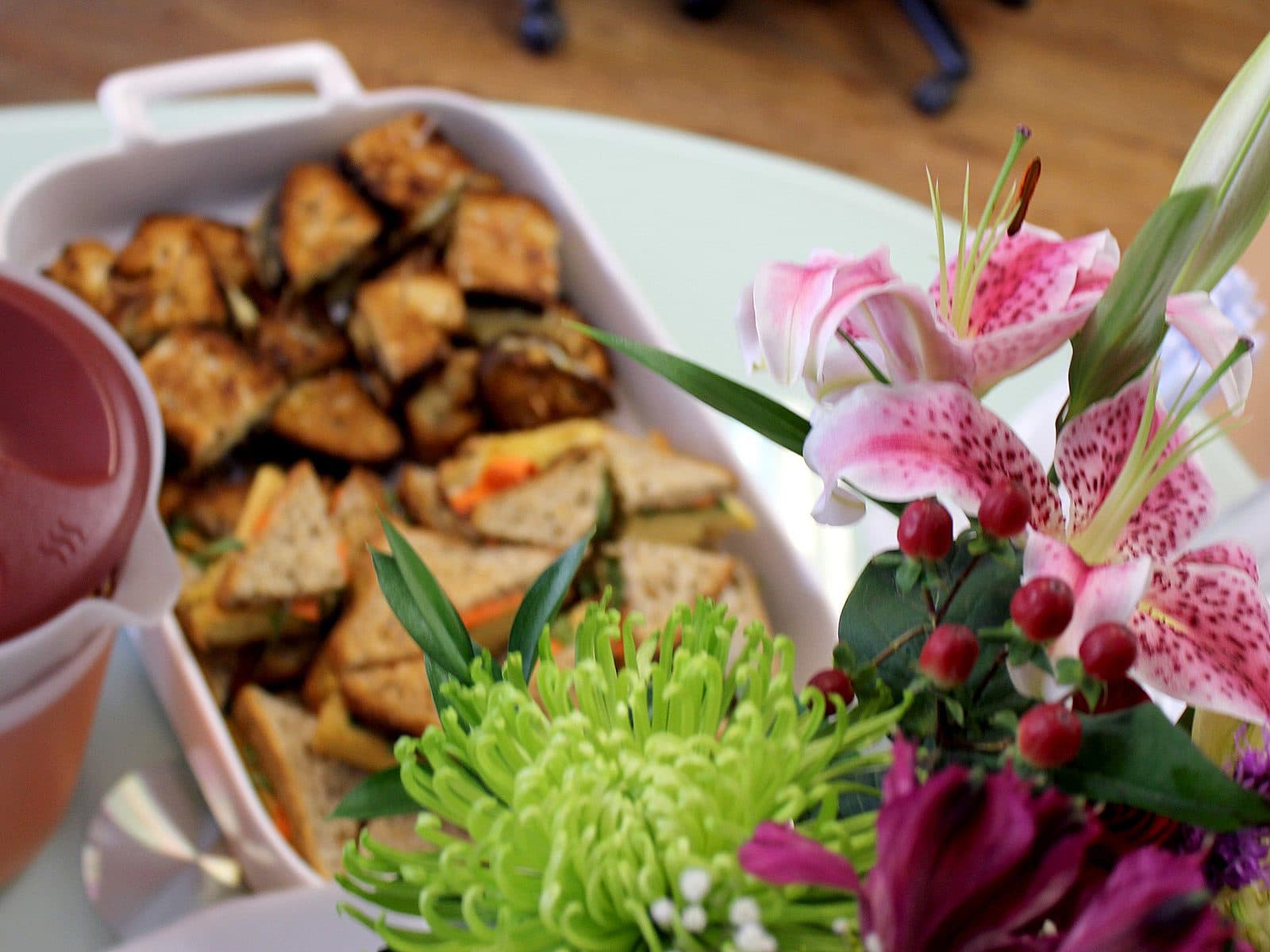A close-up of a platter with sandwiches and snacks next to a bouquet of various flowers, including pink lilies and green chrysanthemums. The background shows a blurry indoor setting with a chair and some indistinguishable items.
