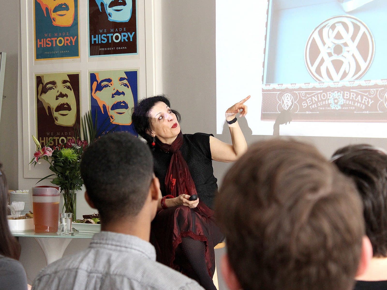 A woman gestures and speaks in front of an audience during a presentation. Behind her is a screen displaying an image and a wall with posters. The audience members are seated, and a table with flowers, a pitcher, and other items is visible.
