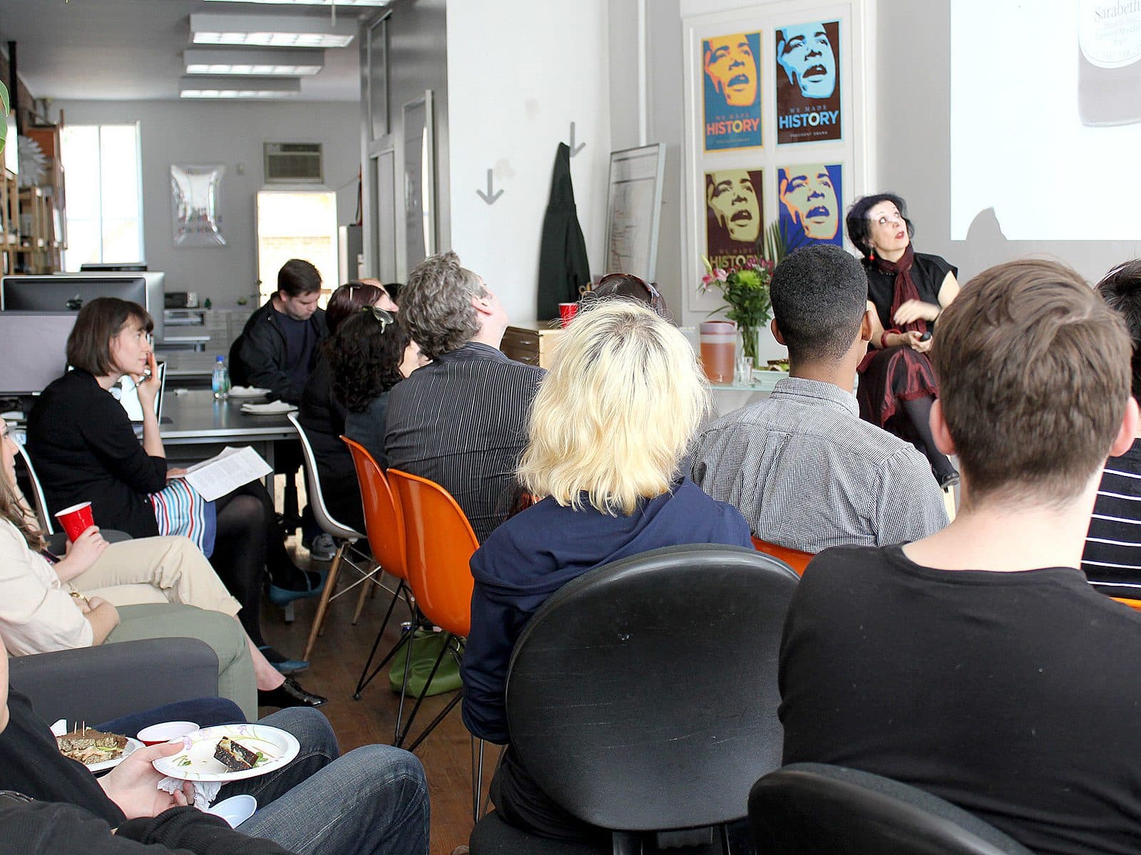 A group of people sit and listen attentively to a presenter in an office setting. The presenter stands near a screen, gesturing as they speak. The room has posters on the wall, a table with snacks, and various office supplies around. Some people hold notebooks and plates of food.