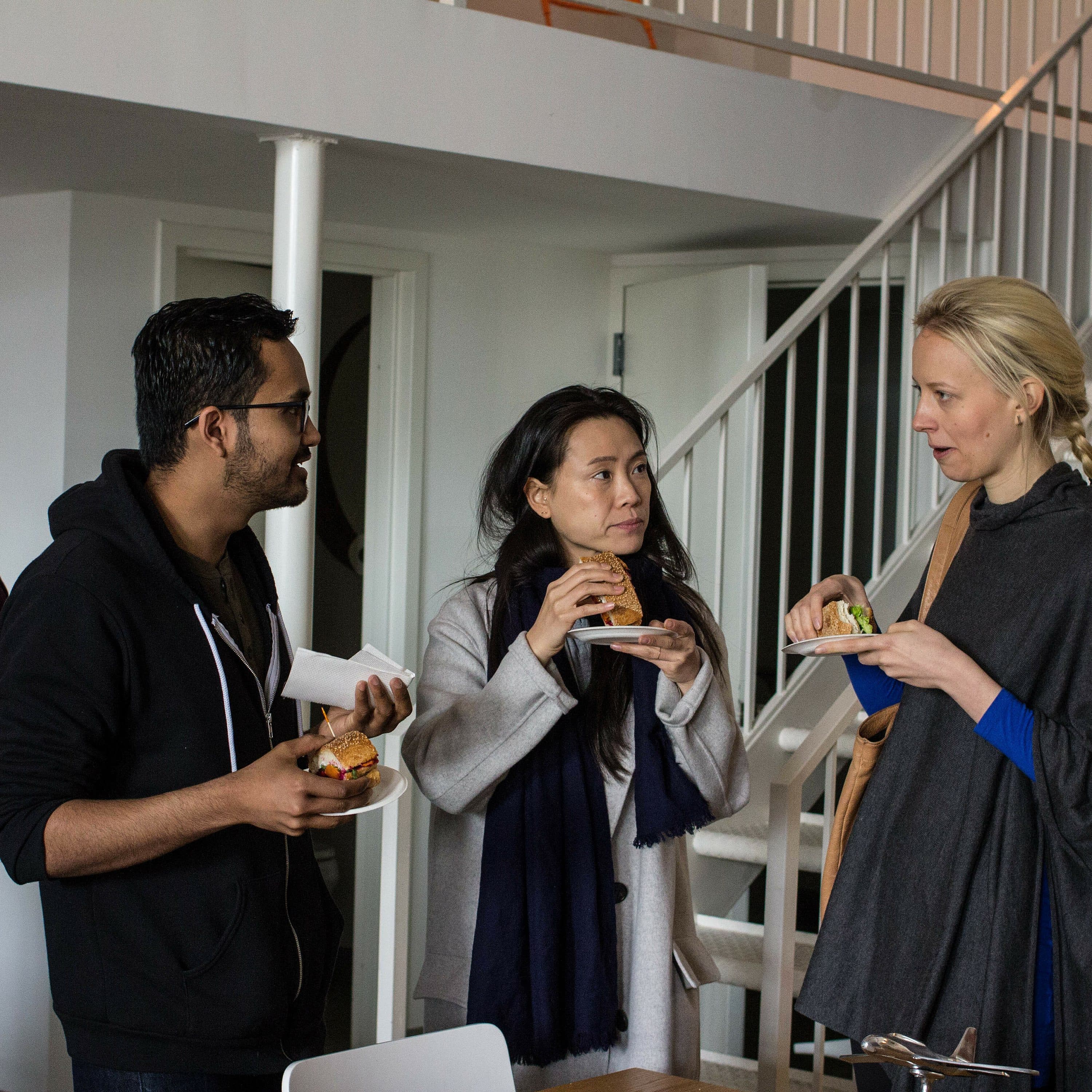Four people are in an indoor setting, engaged in conversation while holding food and drinks. Three are gathered together, eating and talking, while one person in the background looks on. The environment appears casual and social.