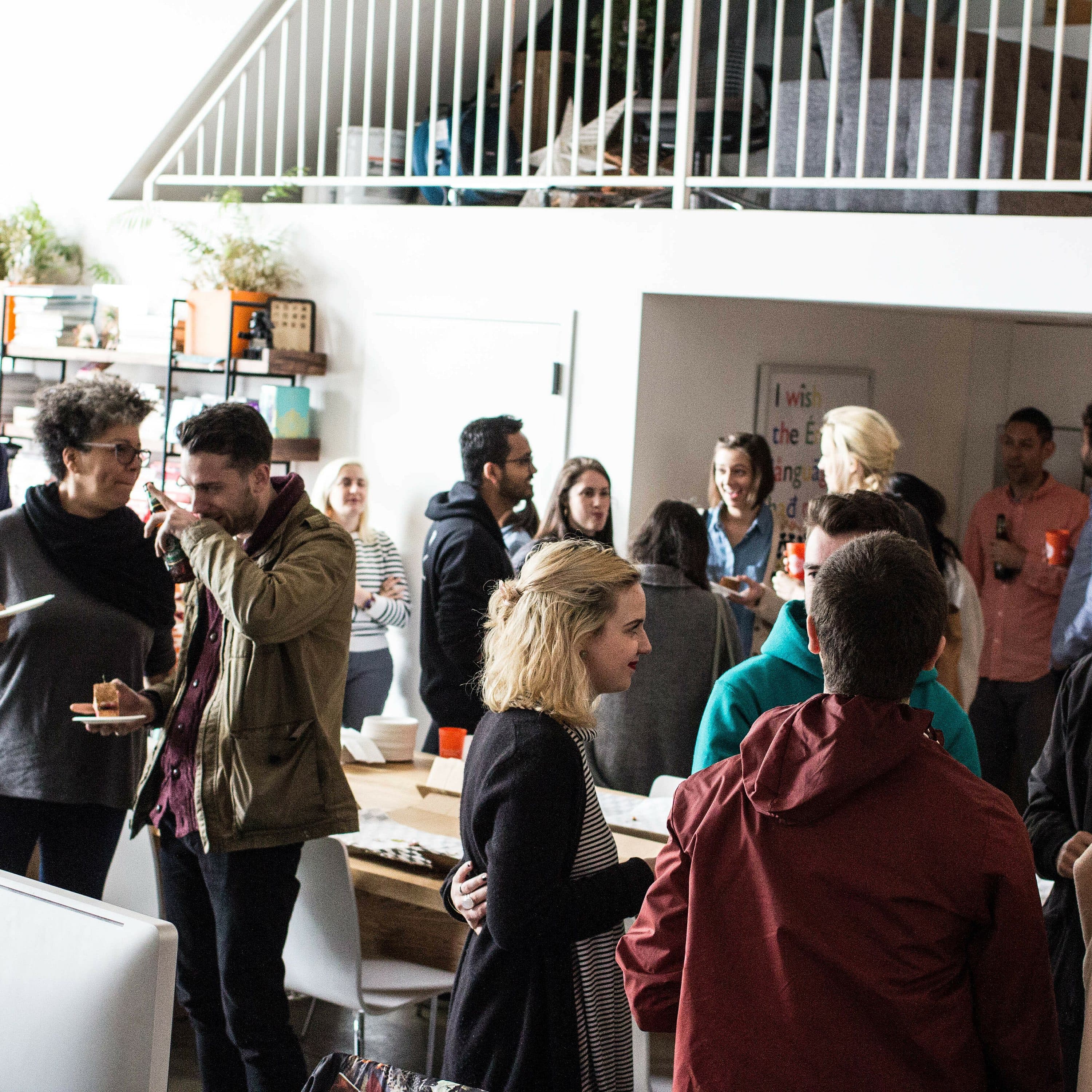 A group of people are socializing at an indoor gathering. Some are standing and talking, while others hold beverages and food. The room is bright with large windows and a loft area above. There are plants and various office items in the background.