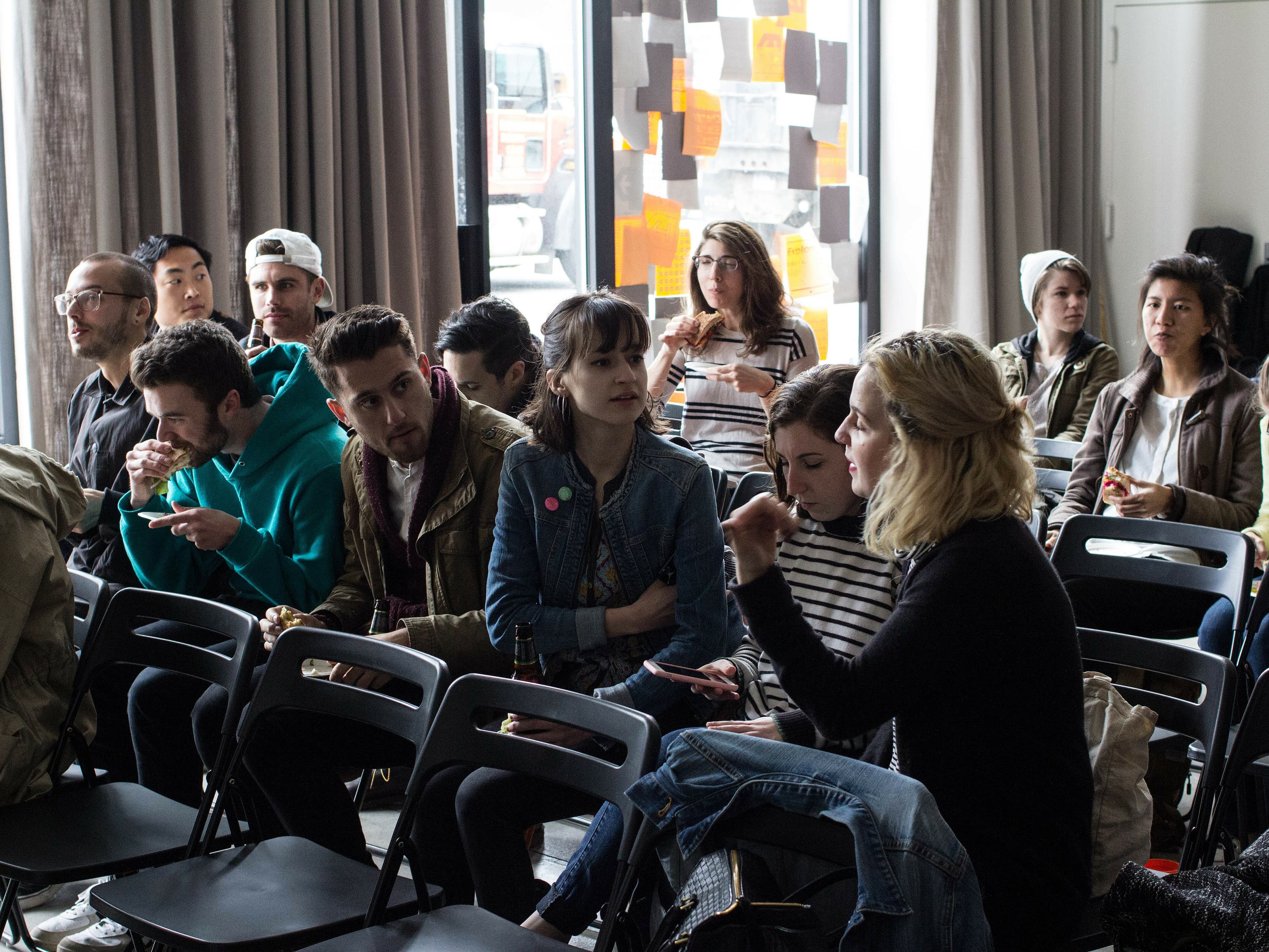 A group of people sits in rows of black chairs in a room, some engaged in conversation while others eat and look at their phones. The room has large windows with abstract decorations. The atmosphere appears casual and relaxed.
