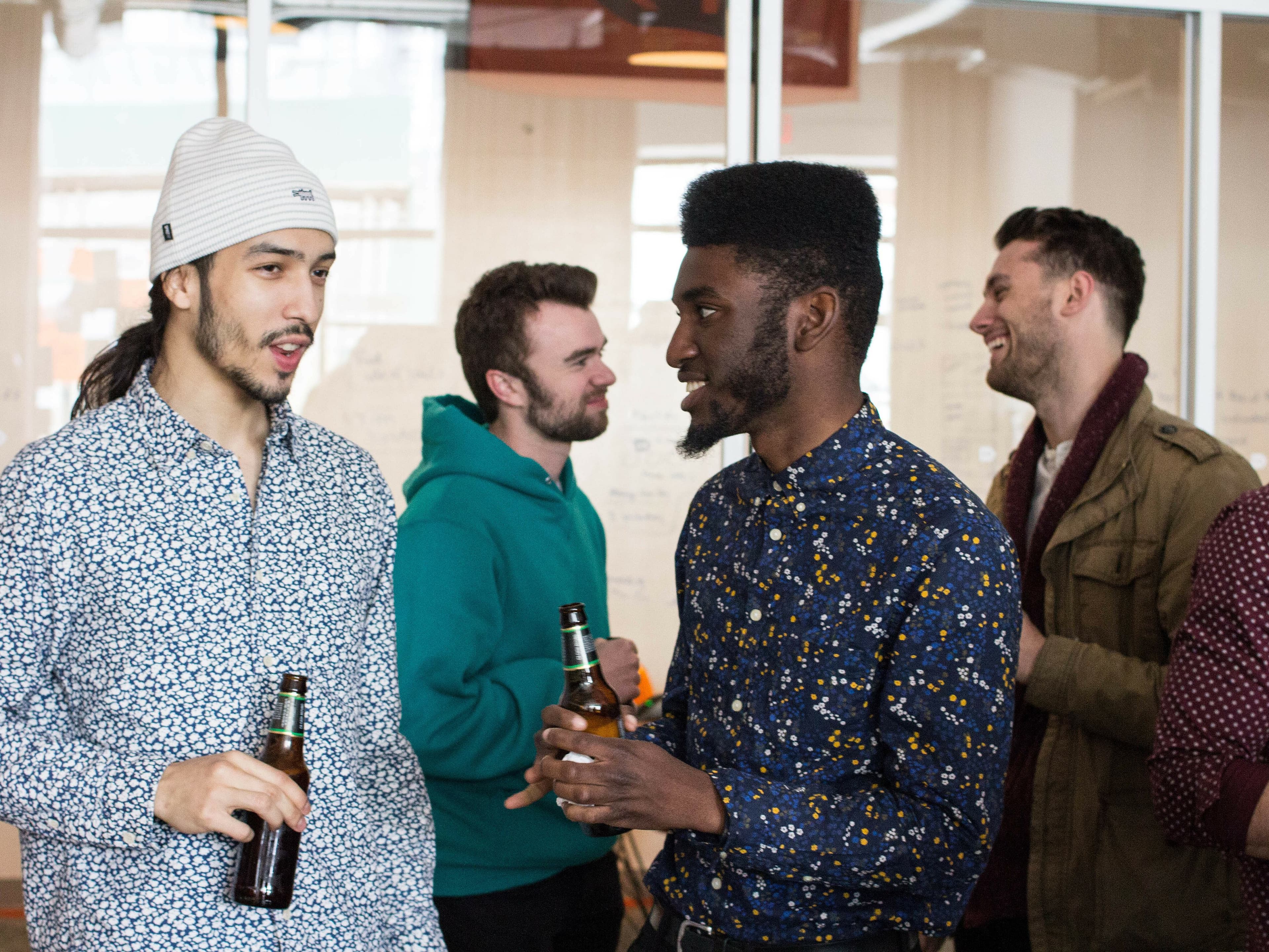 A group of four men standing and talking indoors, each holding a bottle of beer. The men are engaged in conversation and appear relaxed. They are dressed in casual clothing, and the background shows a glass wall with writing, suggesting an informal gathering or event.
