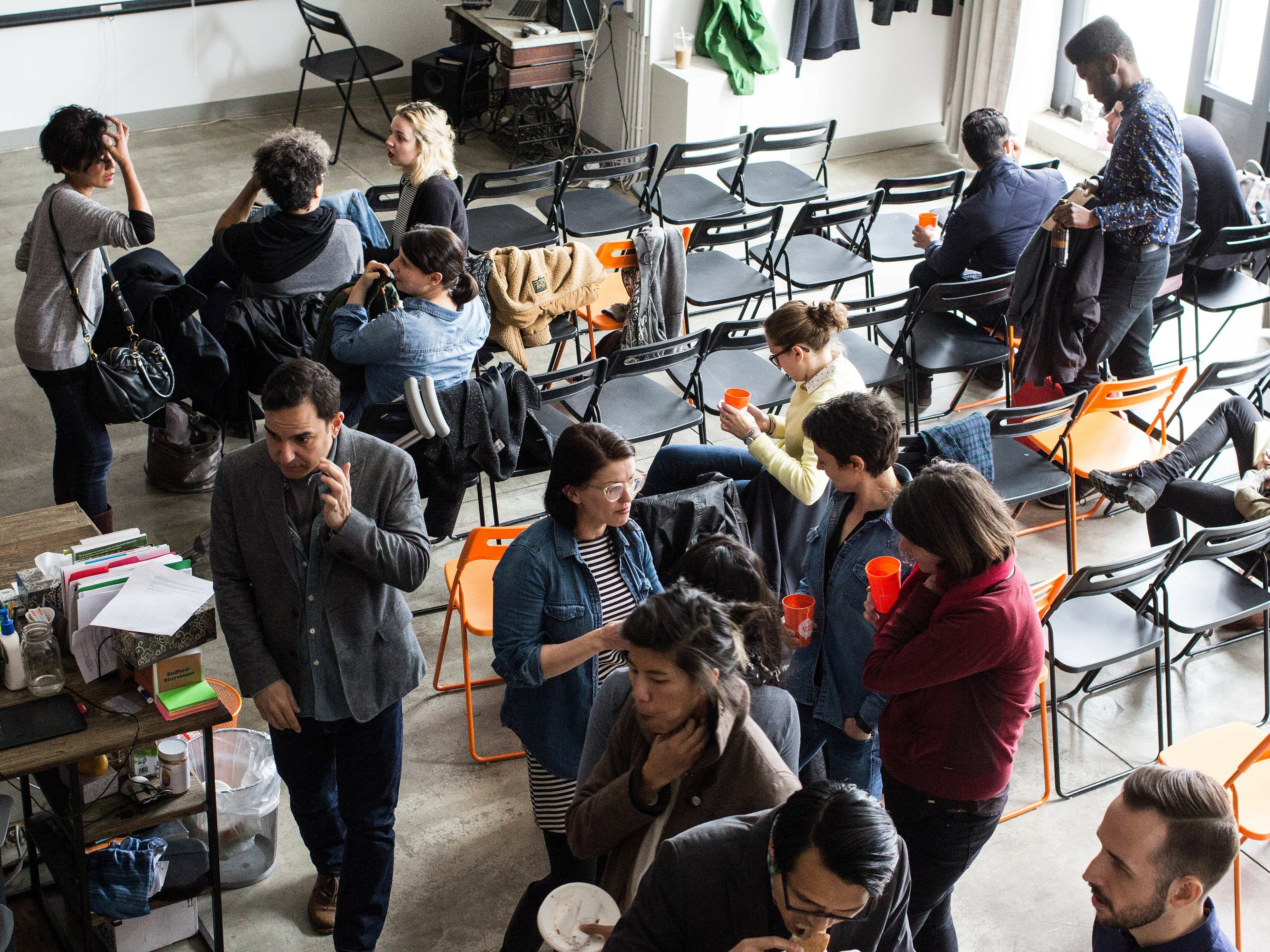 A group of people in a casual setting with scattered chairs, engaging in conversations and using phones. Some are holding drinks, and a few chairs have jackets draped over them. The room has a relaxed atmosphere with a mix of standing and seated individuals.