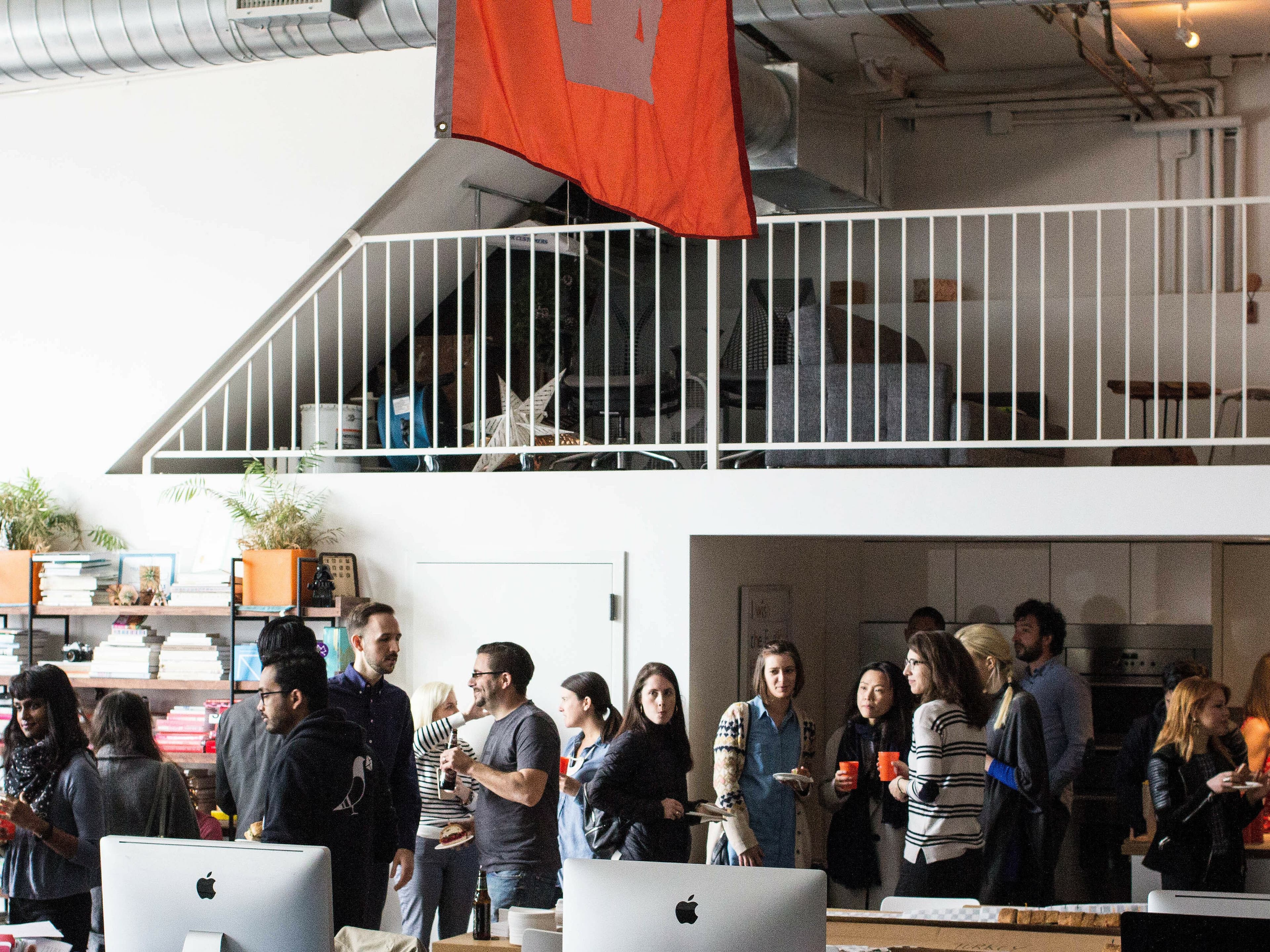People are gathered in a modern office space for a social event. Some are conversing in small groups, while others hold drinks. The room is decorated with bookshelves, plants, and computers. A mezzanine level with a red flag and a star design is visible above.