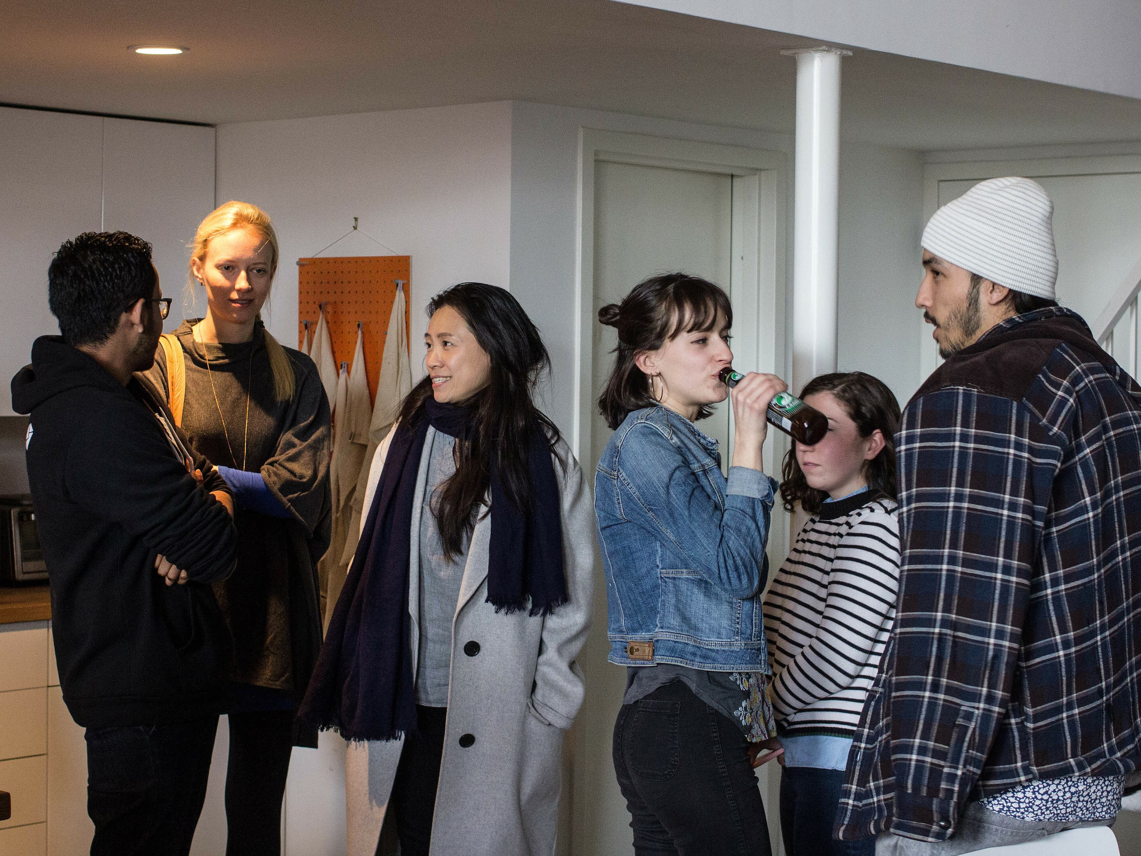 A group of six people stand in a kitchen, engaging in conversation. Two are standing by a countertop on the left, while others are scattered around the space, some talking and one person drinking from a can. A staircase is visible in the background on the right.