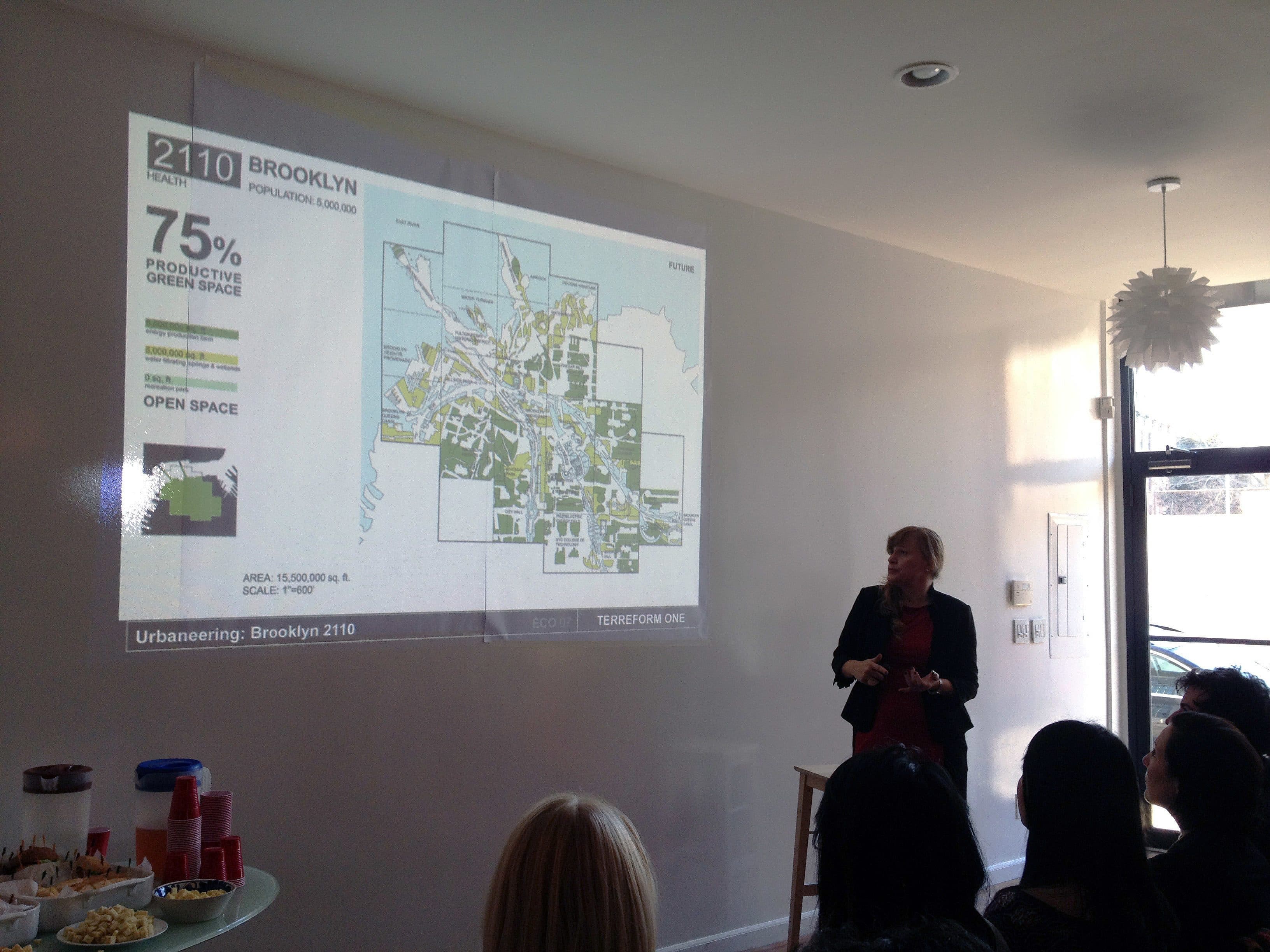 A woman in a dark suit presents a slide at a meeting. The slide is titled "BROOKLYN 2110" and shows a map with a focus on productive green space, marked as 75%. Several people sit watching, and there are refreshments on a nearby table.