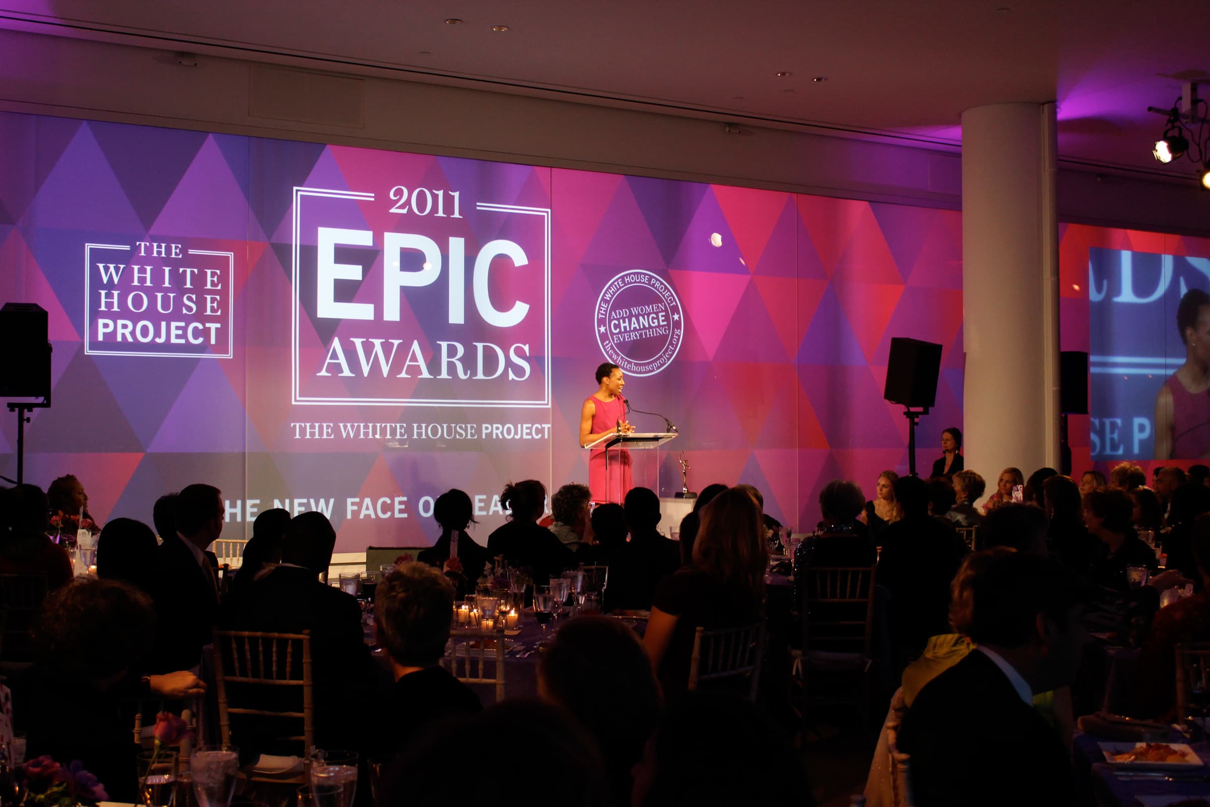 A speaker stands at a podium on a stage at the 2011 EPIC Awards hosted by The White House Project. The audience is seated at round tables, and a backdrop features the event's logos and theme, "The New Face of Leadership." The room is dimly lit with a purple hue.