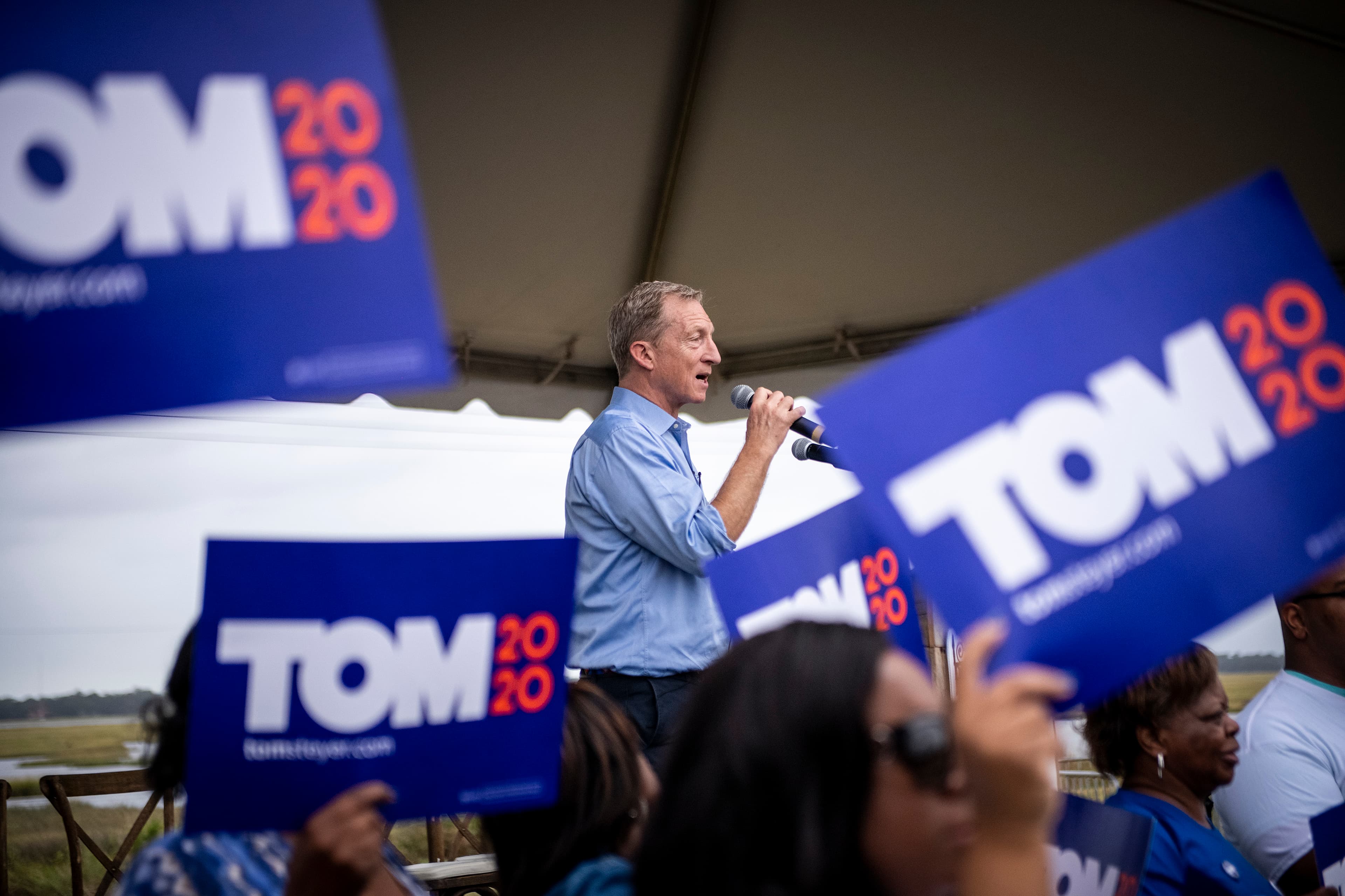 A man speaks into a microphone at a campaign event with supporters holding blue signs that read "TOM 2020." The scene is set under a large canopy, and the man wears a light blue shirt, engaging with an enthusiastic crowd.
