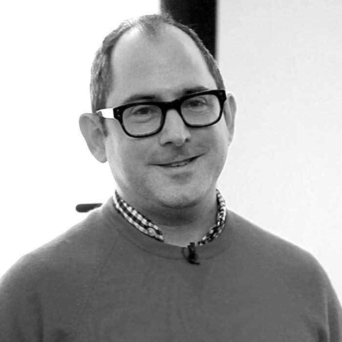 Black and white photo of a man wearing dark-framed glasses, a collared checkered shirt, and a crew-neck sweater. He has short hair and is smiling slightly while looking at the camera. The background is blurred.