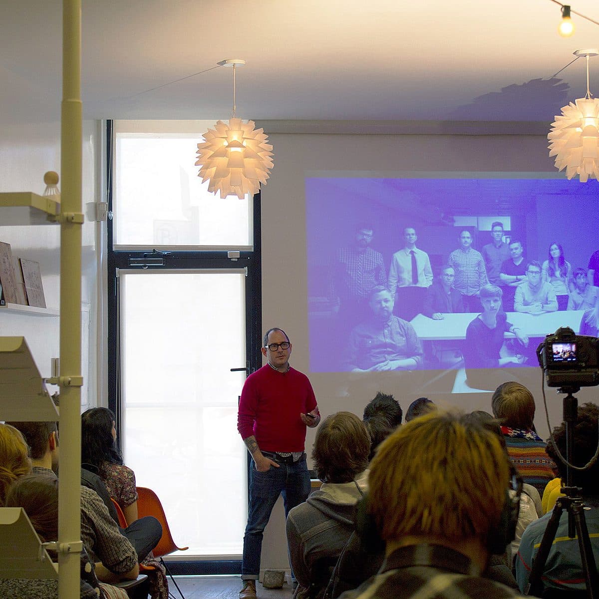 A man in a red shirt stands in front of an audience, giving a presentation. Behind him is a projected image of a group of people posing for a photo. The room is well-lit with large hanging lights and shelves on the left side. A camera is set up recording the event.