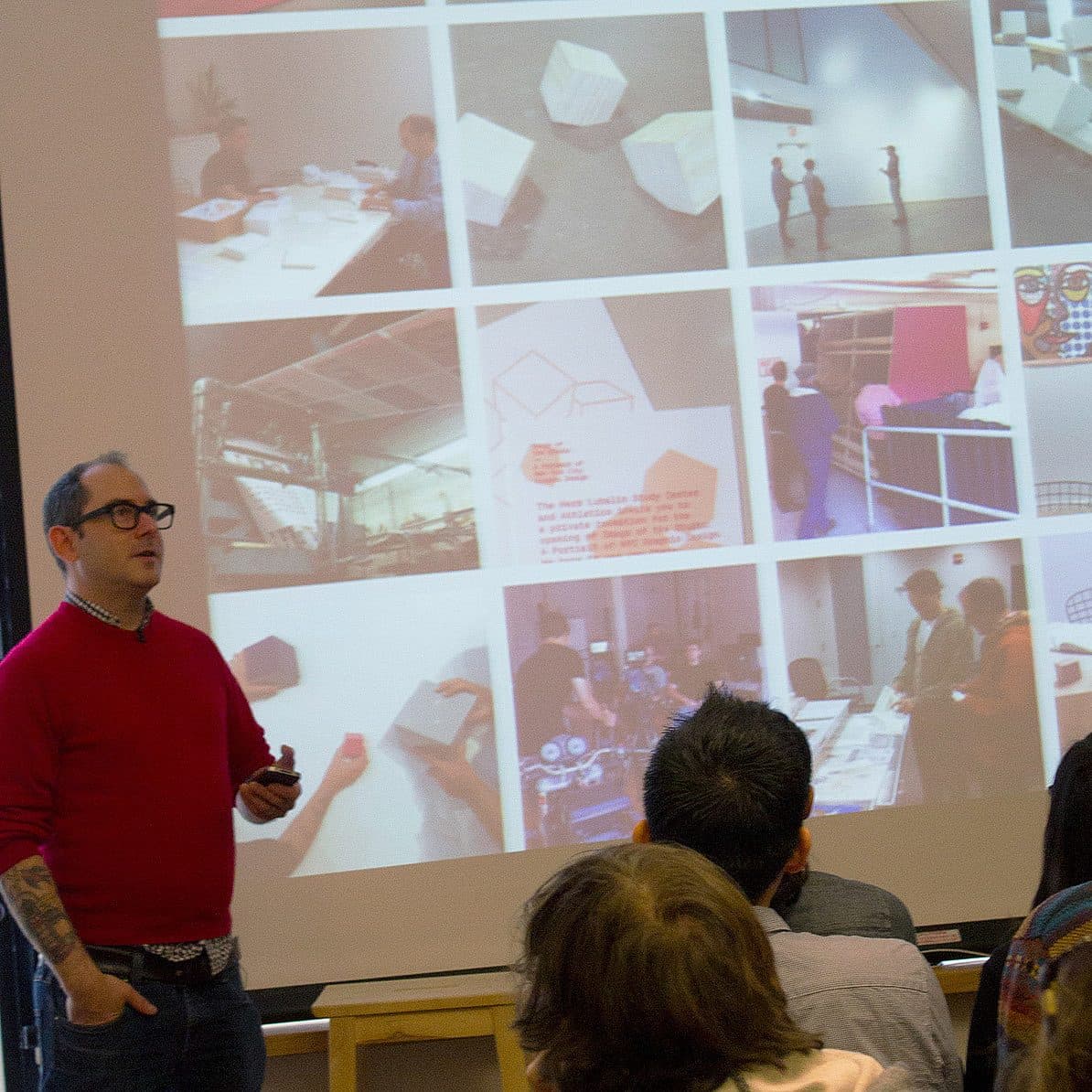 A speaker in a red sweater and glasses stands in front of an audience, presenting a slideshow featuring various images of design and architecture projects. The audience is seated and attentively watching the presentation.