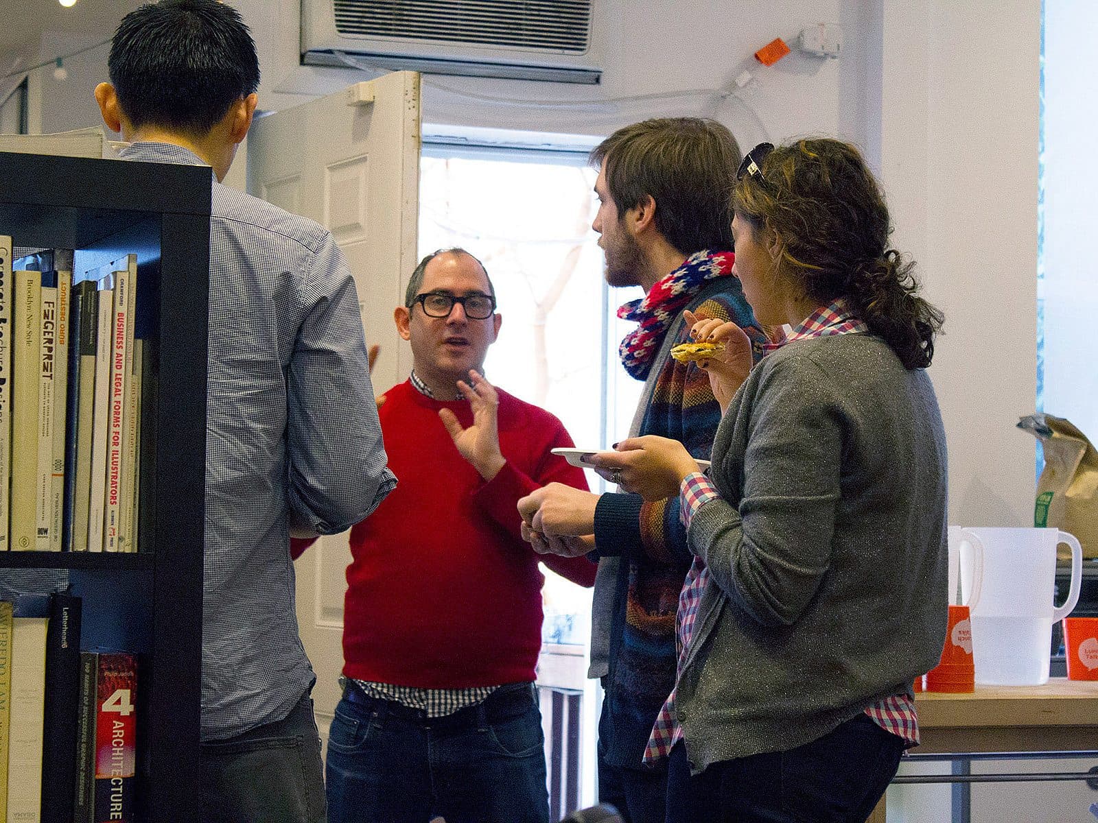 Four people are engaged in conversation in an office space. Three are standing near a bookshelf while one is at a table with food and cups. A man in a red sweater gestures as he speaks, while the others listen and hold drinks and plates of food.