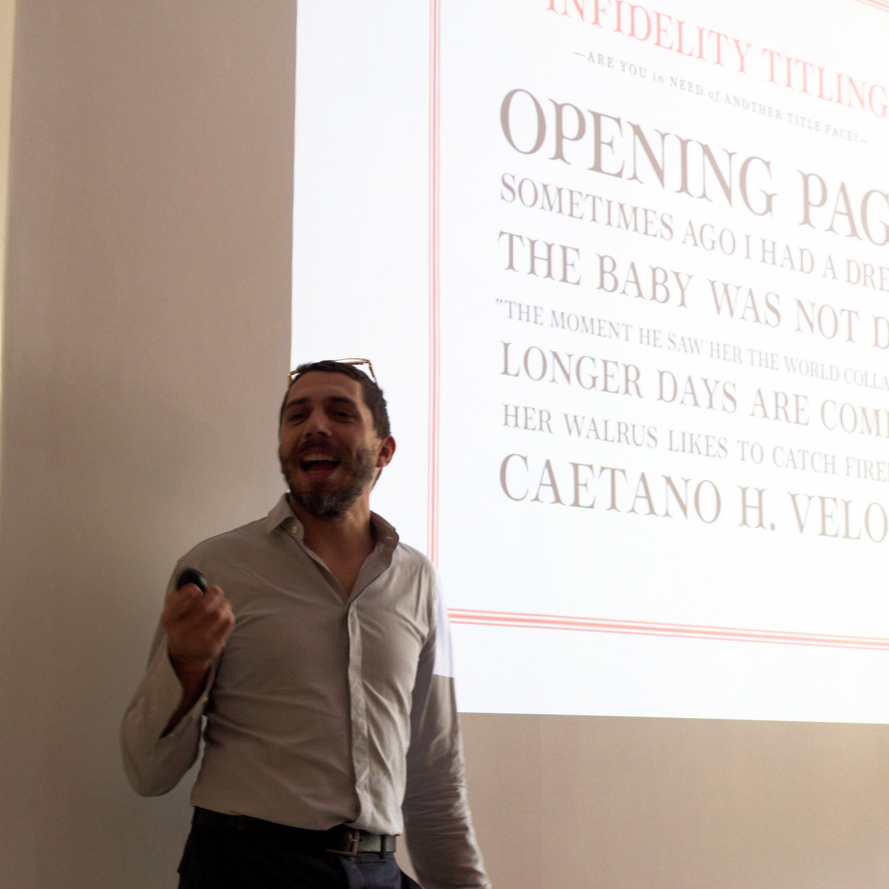 A man wearing glasses and a striped shirt is giving a presentation. He is holding a remote in one hand and is smiling. The projected slide behind him features a mock book cover with the title "Infidelity Titling" and other sample text.