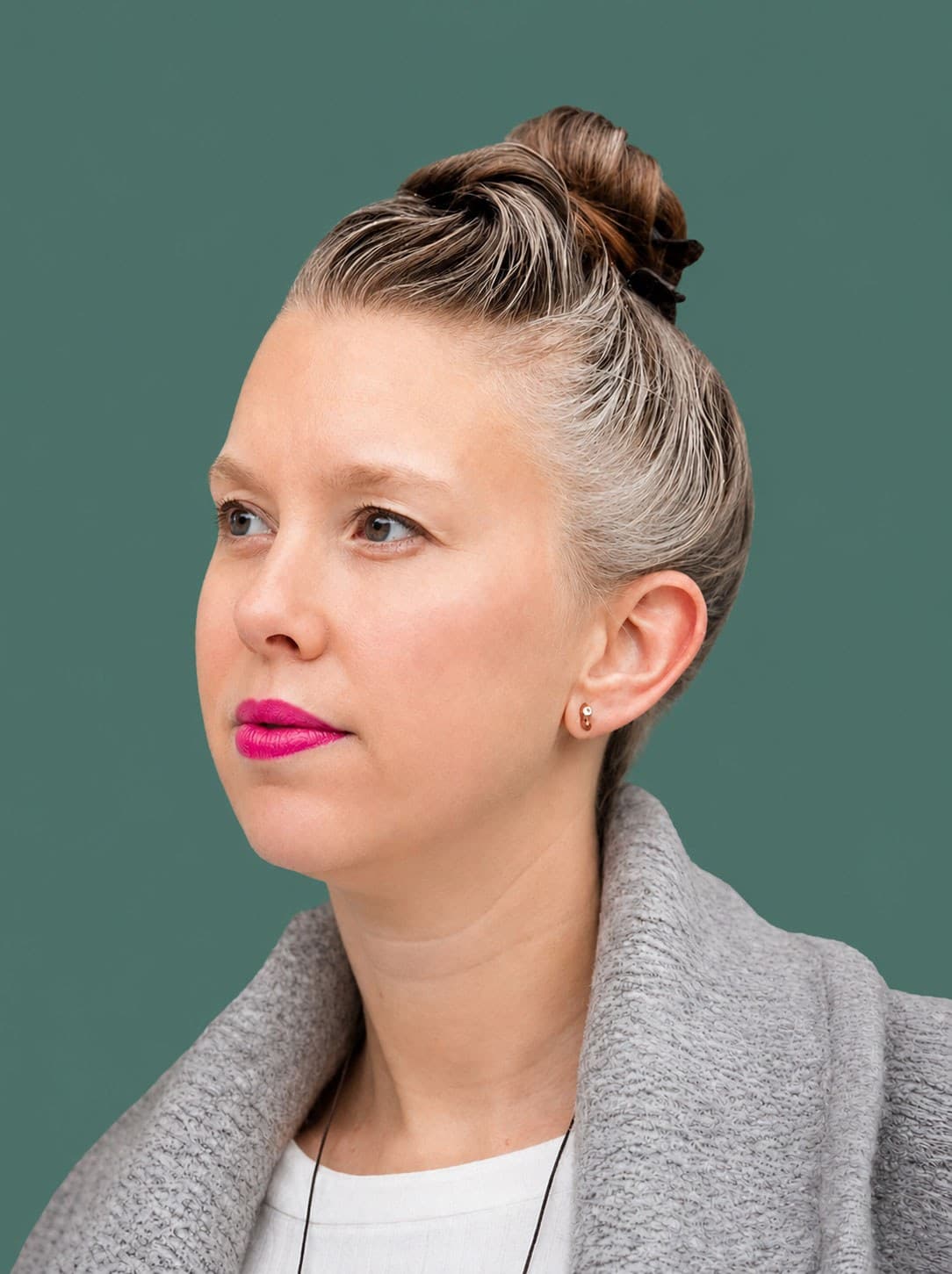 A person with light skin and gray-streaked hair in a high bun is wearing bright pink lipstick, a gray coat, and a white top, looking slightly to the left against a plain green background.
