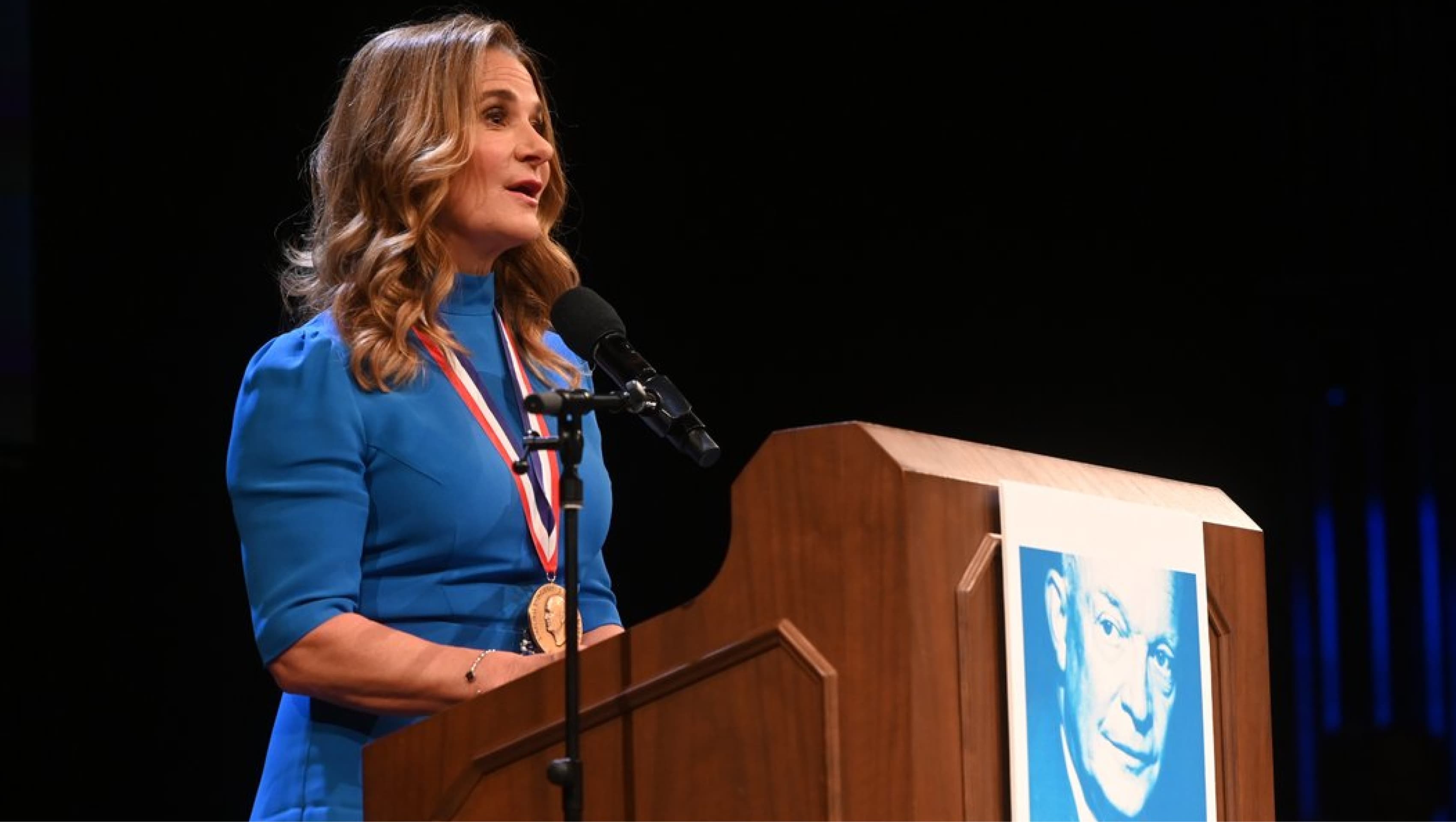 A woman in a blue dress with a medal around her neck speaks at a podium with a microphone. The podium has a large blue-and-white portrait of a man attached to the front.
