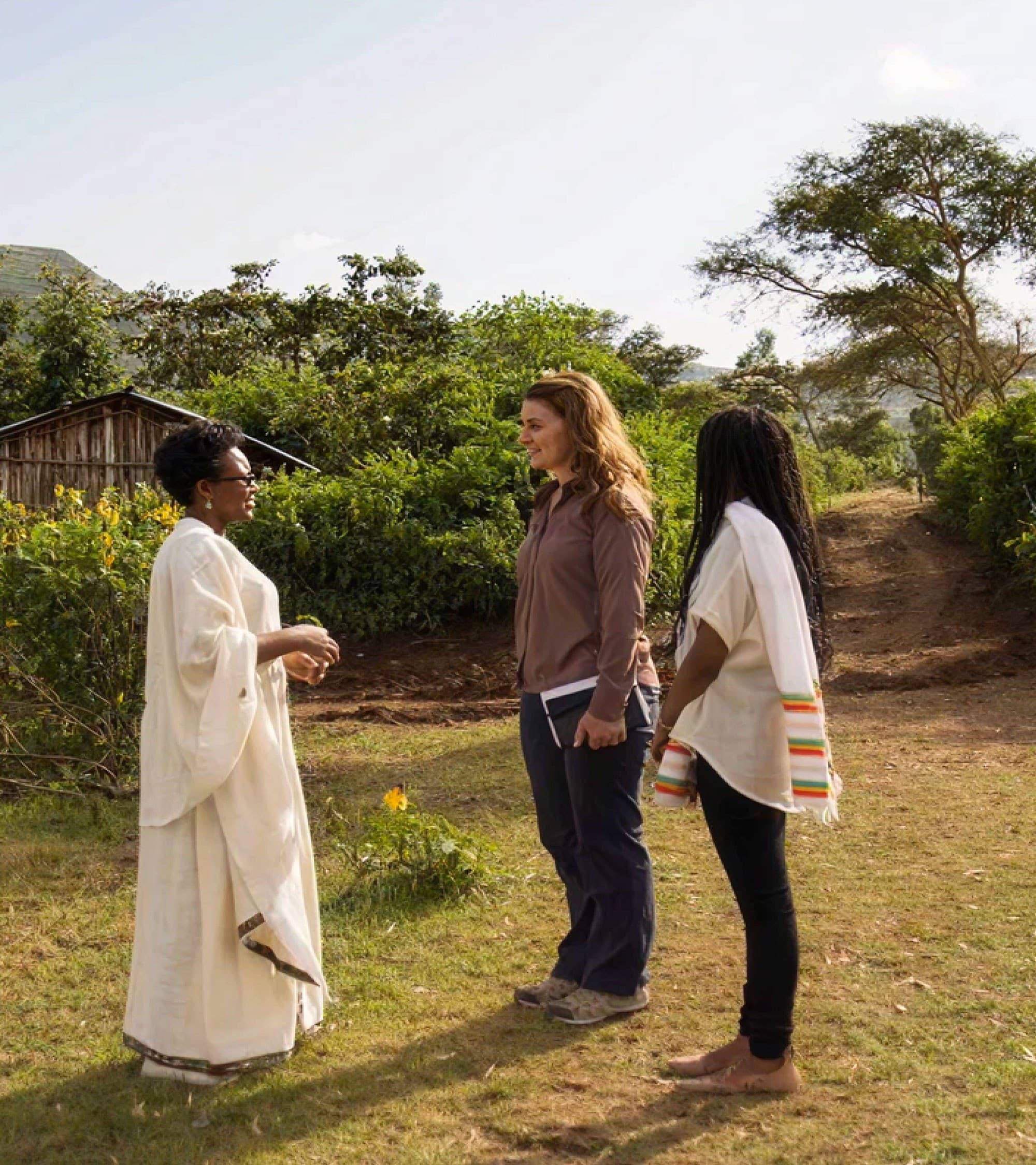 Three women stand outside talking on a dirt path near a rural house, surrounded by greenery and trees. One woman wears a white traditional robe, while the other two wear casual clothes. The sky is clear and sunny.