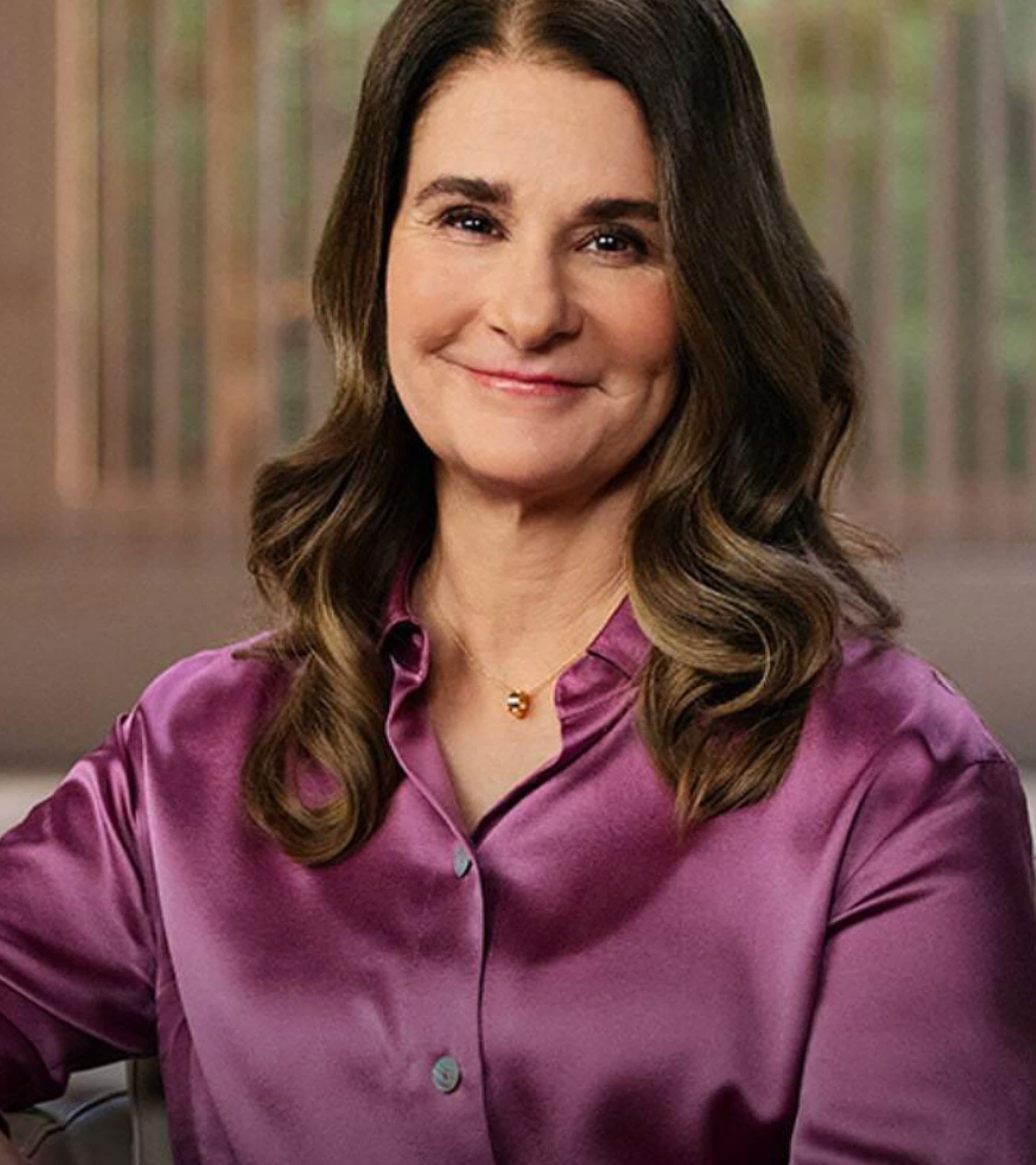 A woman with long brown, wavy hair wearing a silky purple blouse smiles gently while sitting indoors, with a blurred background featuring wooden elements.