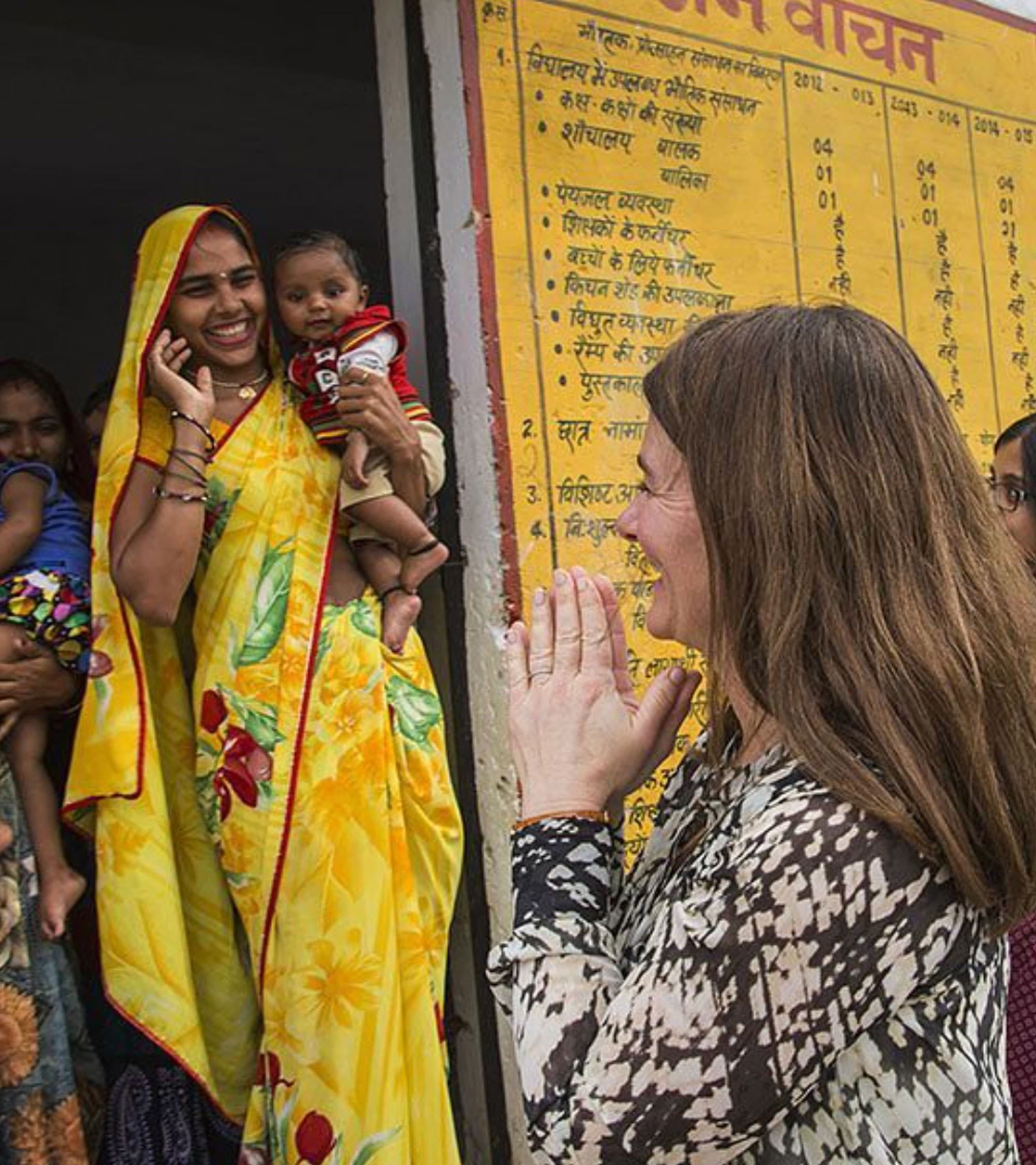 A woman in a yellow sari holding a baby smiles while greeting another woman who has her hands together in a respectful gesture; several people and a yellow poster with Hindi text are in the background.