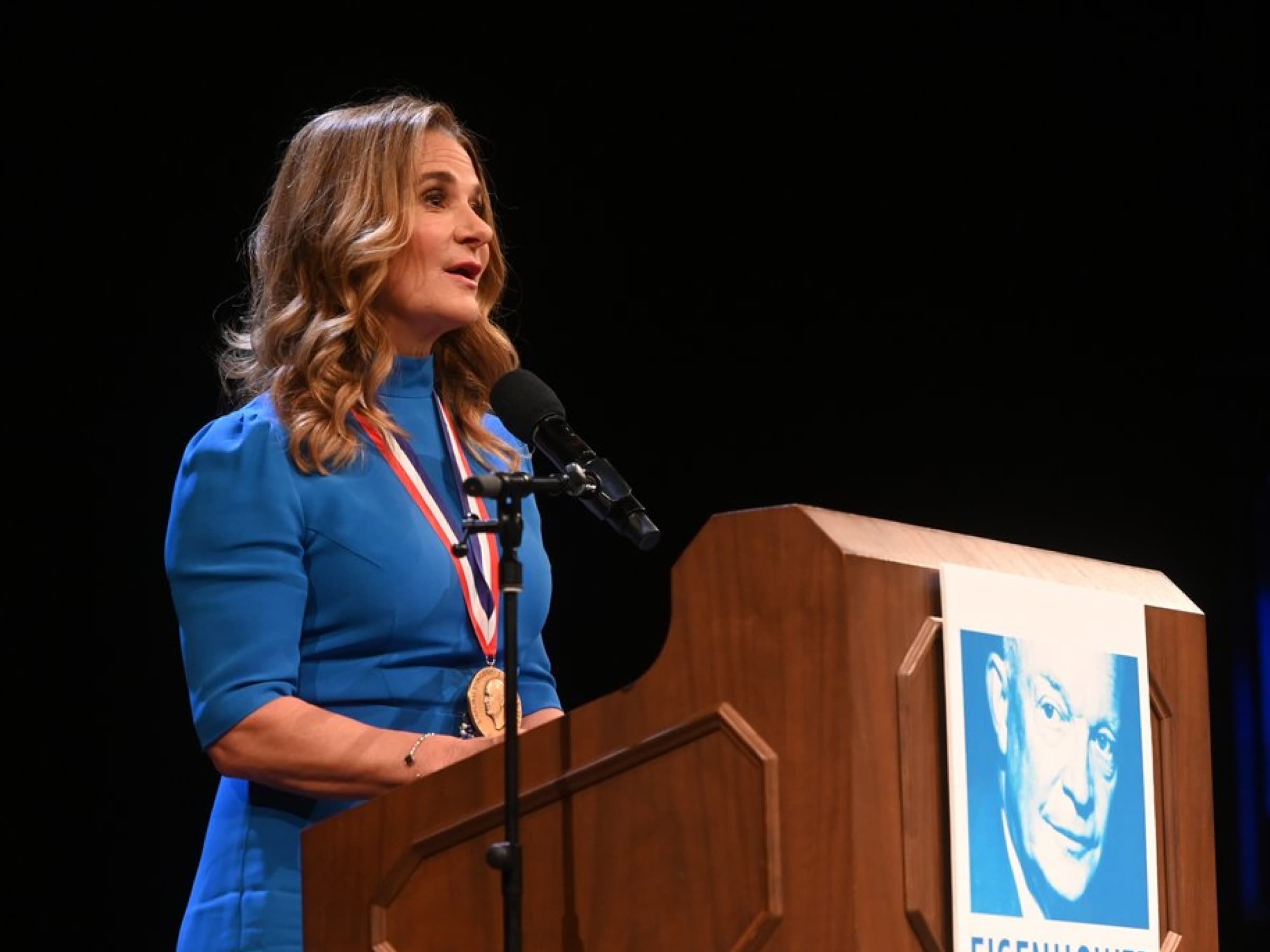 A woman in a blue dress wearing a medal speaks into a microphone at a wooden podium with a photo of Dwight D. Eisenhower attached to the front.