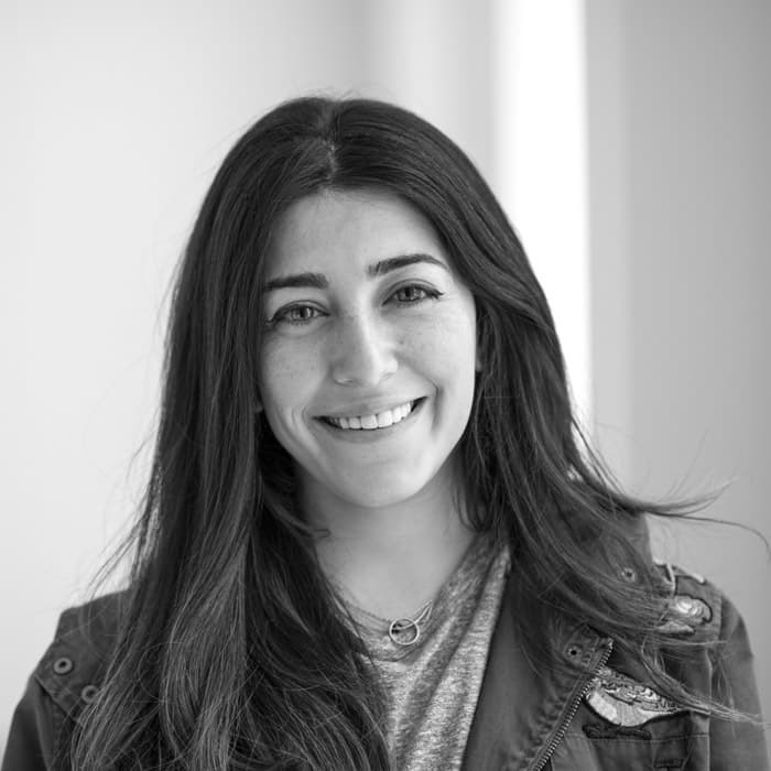 Black and white photo of a woman with long, dark hair smiling at the camera. She is wearing a light-colored shirt and jacket with a circular necklace. The background appears softly lit.