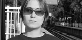 A person with short hair and wearing sunglasses stands in front of a train platform. The image is in black and white. The individual looks directly at the camera with a neutral expression. The background includes a railway track and some trees.
