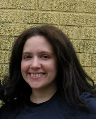 A woman with long, dark hair is smiling in front of a yellow brick wall. She is wearing a dark top.