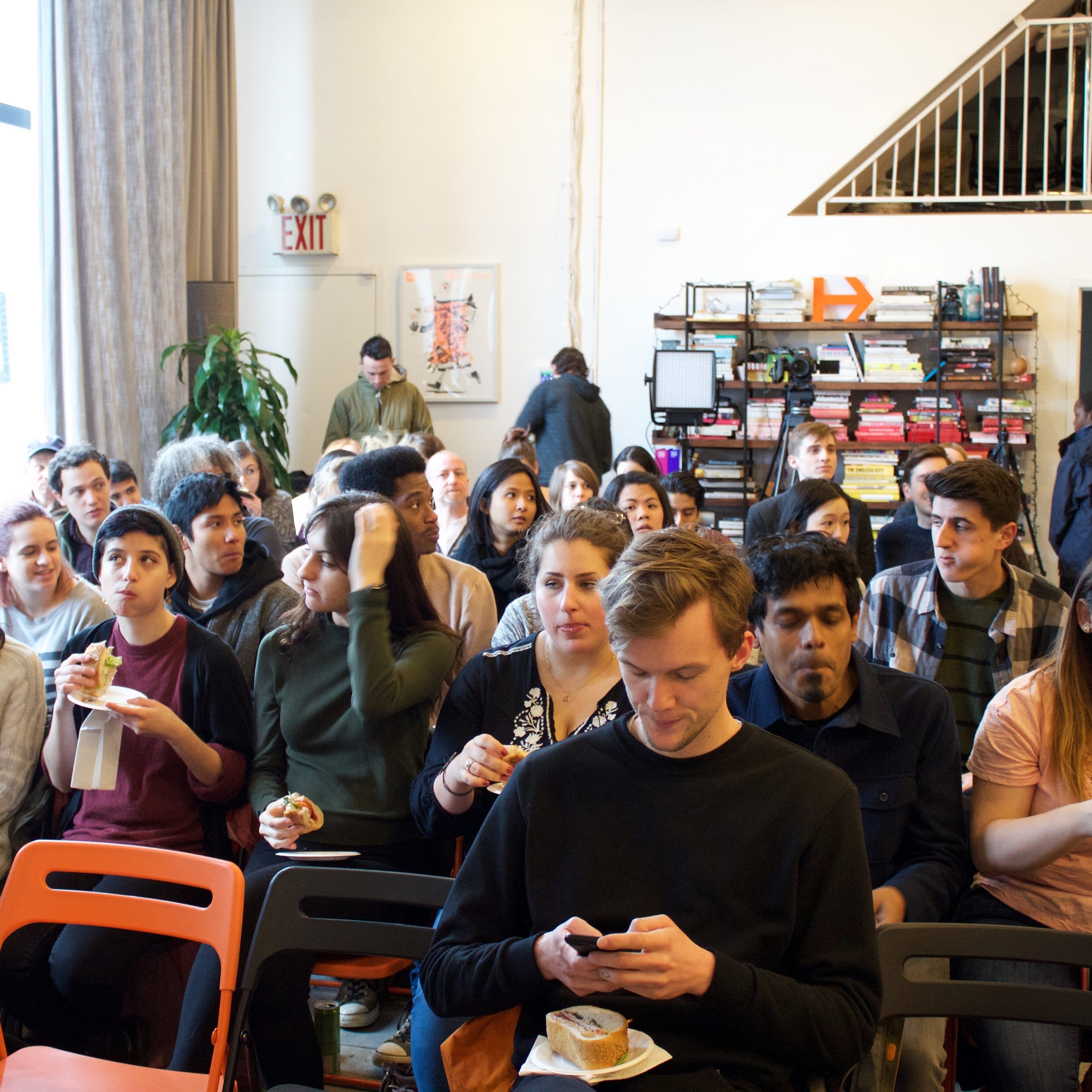A group of people sit in rows of chairs, facing forward in a well-lit room with large windows to the left. Some are eating while others are looking at their phones or talking. Shelves with books and items are against the wall, and a staircase leads to an upper level.