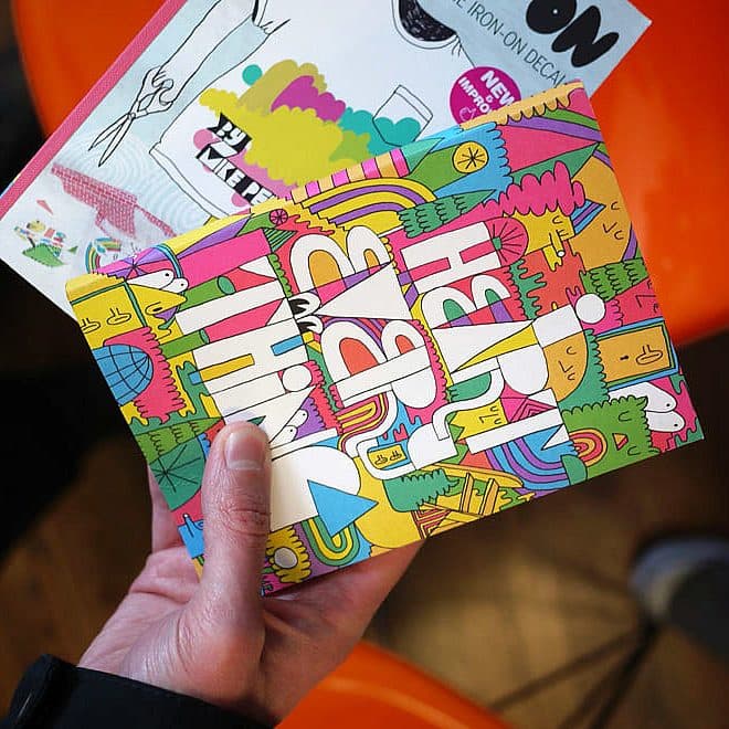 A hand holds a colorful, illustrated brochure with vibrant, abstract designs and text. Part of another similar brochure is visible on top. The background shows an orange chair and part of a wooden floor.