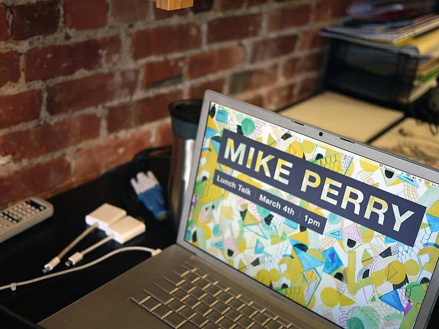 A laptop on a desk displays a colorful screen promoting a "Mike Perry Lunch Talk" scheduled for March 4th at 1 pm. The desk also has a calculator, some cables, and a coffee tumbler, with a brick wall and organized documents in the background.