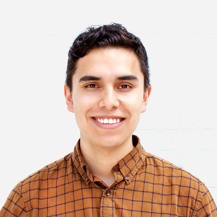 A person with short, dark hair is smiling at the camera. They are wearing a brown checkered button-up shirt and are standing against a plain white background.