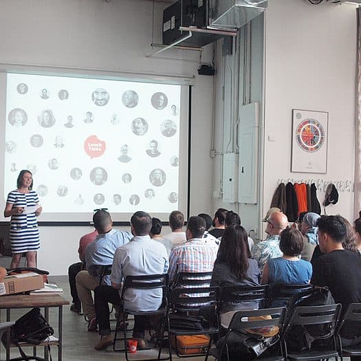 A speaker stands in front of a group of seated attendees in a well-lit room, presenting a slideshow projected on a screen. The slide features a large red circle with text and various smaller black-and-white circles with faces inside them.