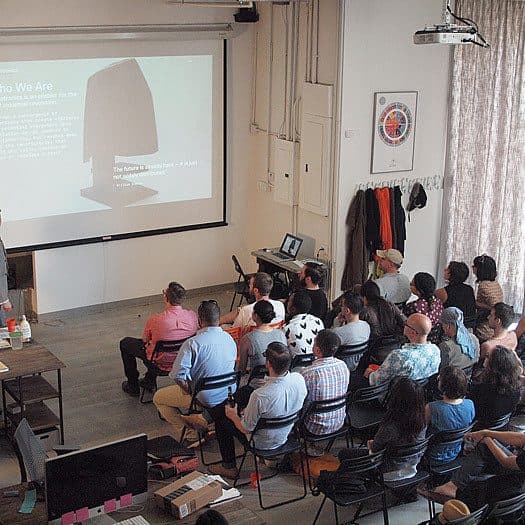 A group of people seated in a room attentively watch a presentation. The presenter stands at the front near a large projected screen that displays text titled "Who We Are" and an image of a hoodie. A few desks with laptops and other materials are in the foreground.