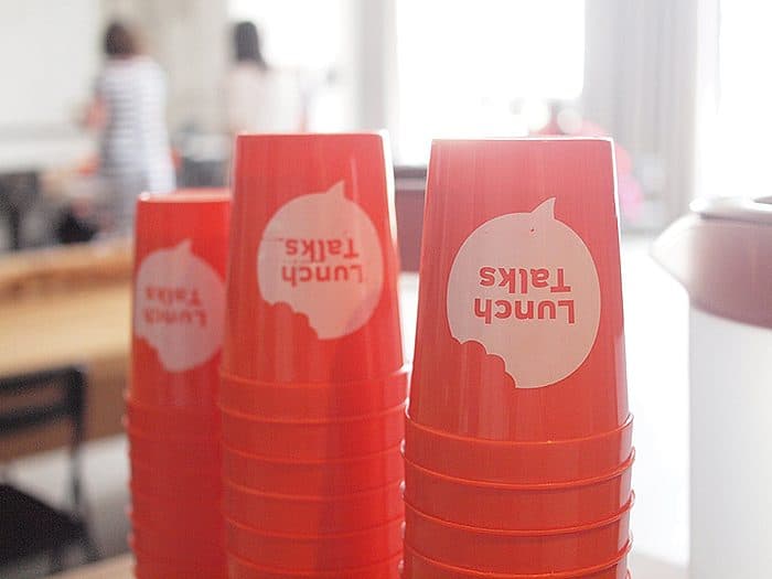 A stack of red cups with the words "Lunch Talks" printed on them in white. The image shows three stacks of cups on a table. Blurry figures of people are seen in the background. The setting appears to be a casual indoor environment.