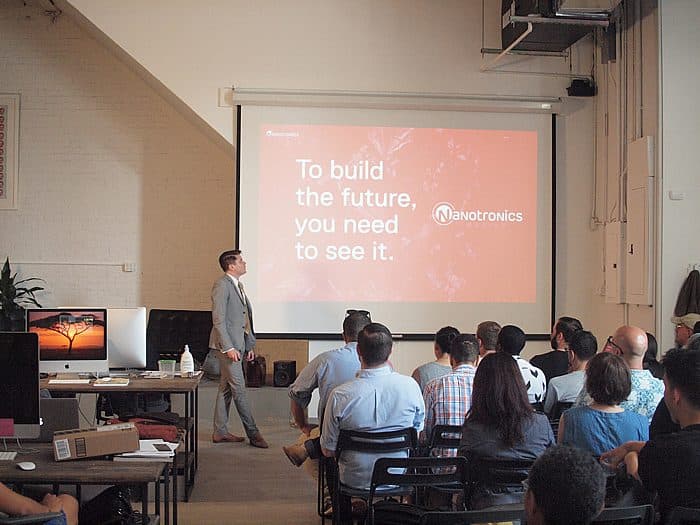 A speaker in a gray suit is presenting to an audience in a bright room. The slide on the screen reads, "To build the future, you need to see it. Nanotronics." The audience is seated and attentively watching the presentation.