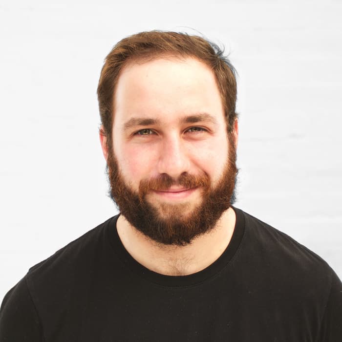 A bearded man with short, dark hair and a slight smile stands against a plain white background. He is wearing a black t-shirt.