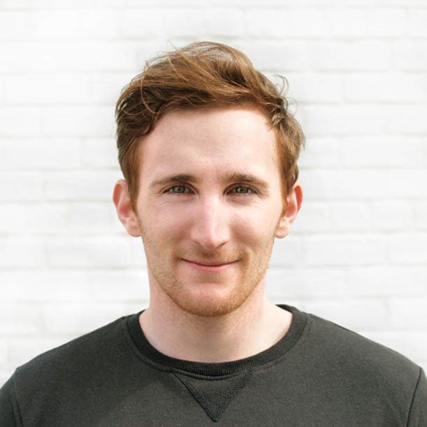 A person with short, reddish-brown hair and a light stubble smiles gently at the camera. They wear a dark crewneck sweater, and the background is a white brick wall.