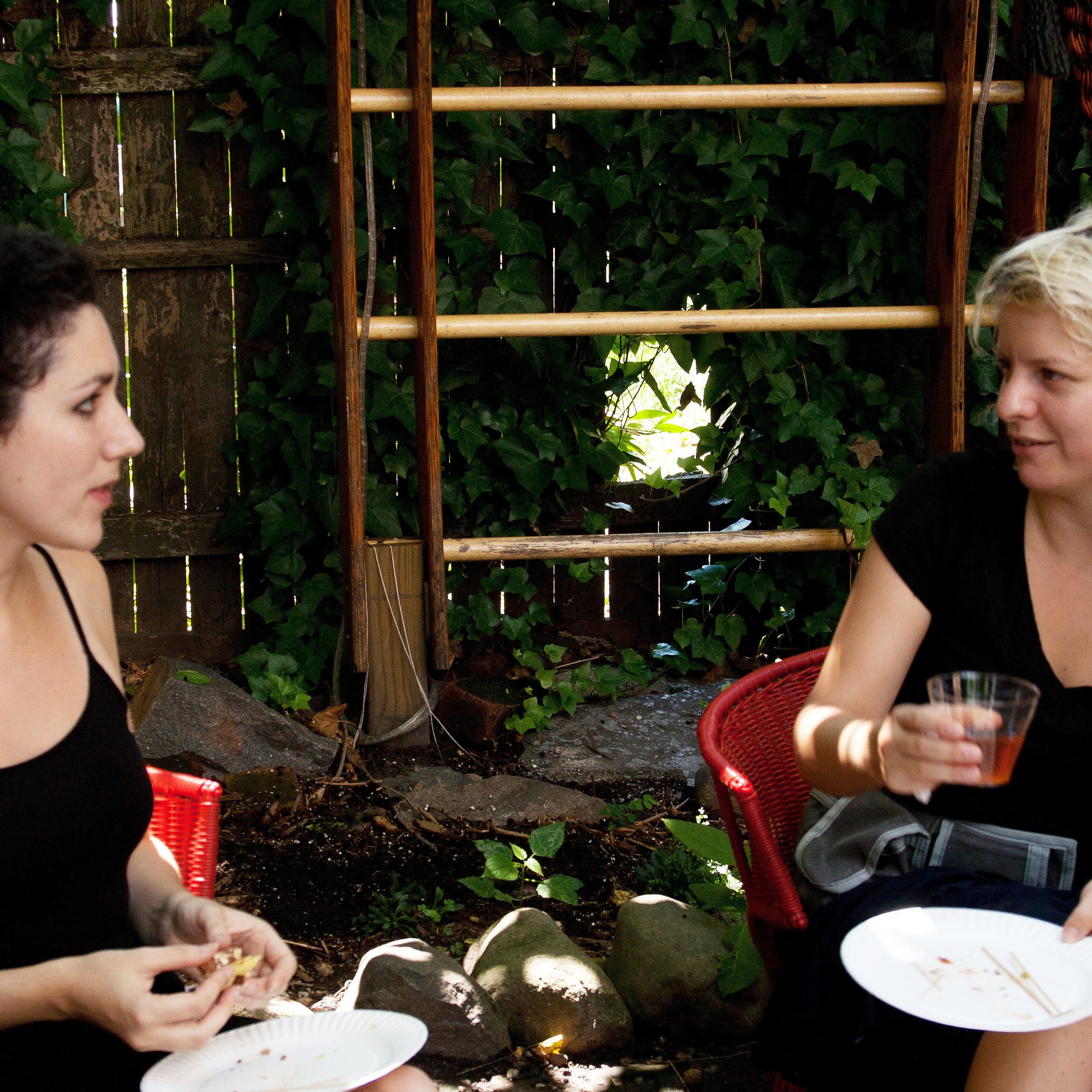 Two women are sitting outside on red chairs, engaged in conversation. Both are holding white disposable plates, with one woman also holding a drink in a clear plastic cup. They are surrounded by greenery and a wooden fence, giving the setting a cozy, garden-like atmosphere.