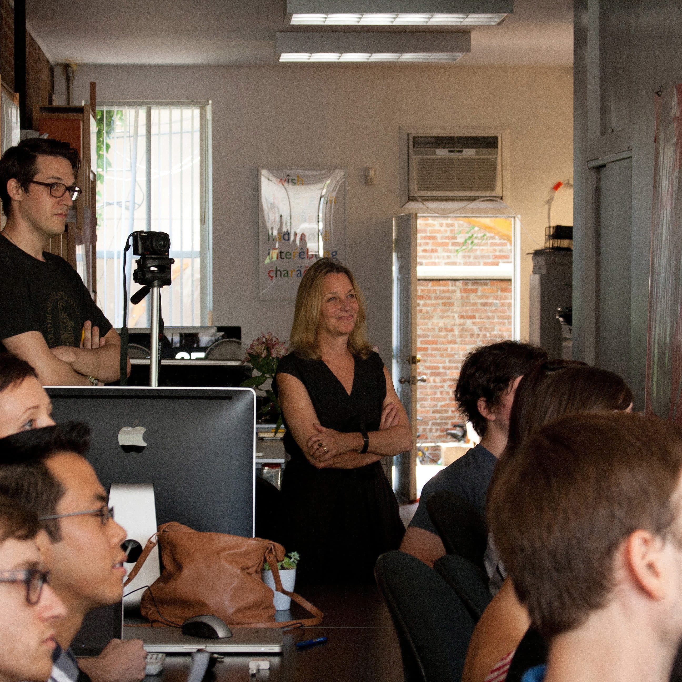 A group of people sitting and standing in an office space, attentively facing towards a presentation or speaker off-frame. A man stands near a camera, and a woman smiles in the background. Various equipment and shelves are visible around the room.