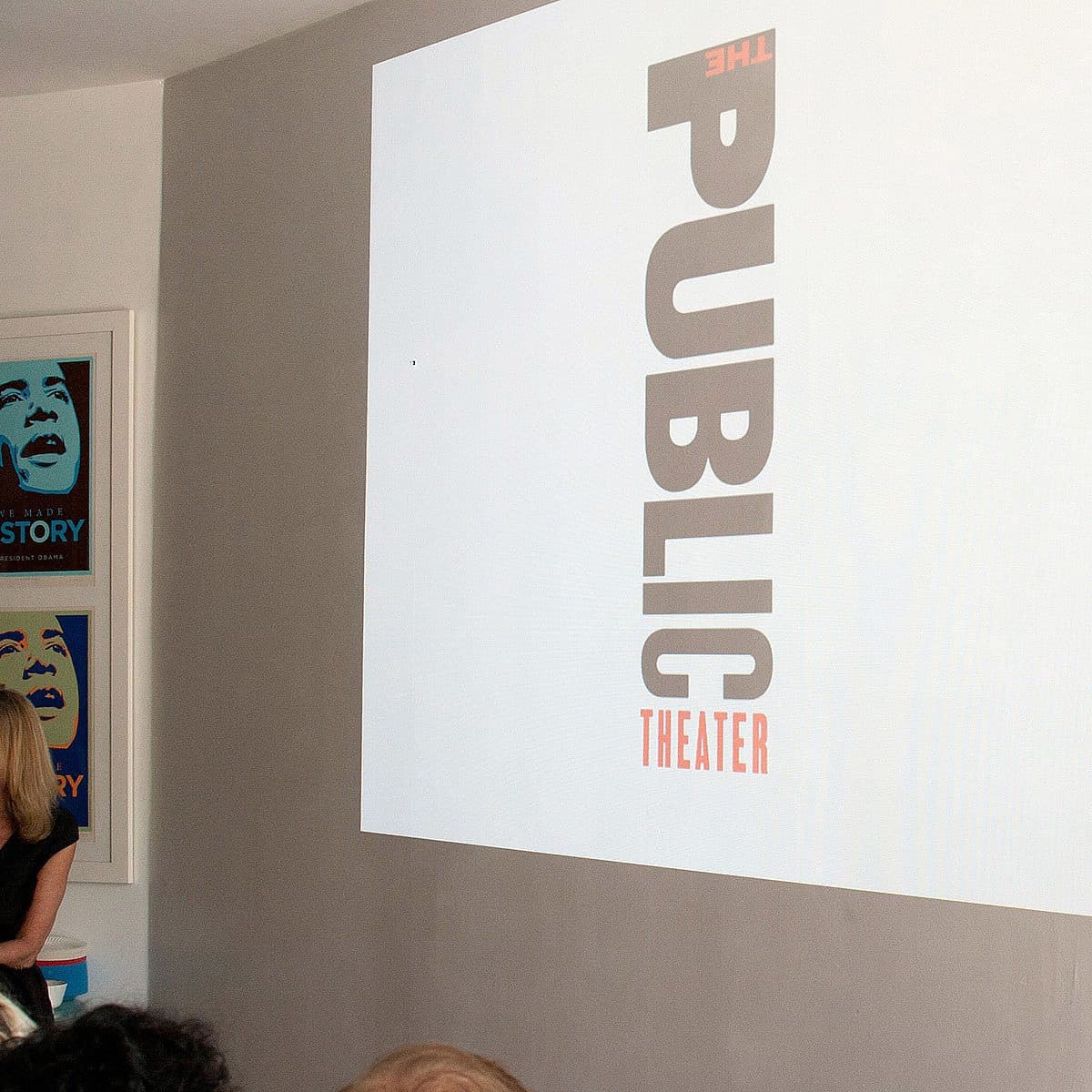 A woman sits on a chair, facing an audience, next to a projection screen displaying the words "The Public Theater" in large, bold letters. Behind her, framed posters featuring stylized portraits and the word "HISTORY" adorn the wall.