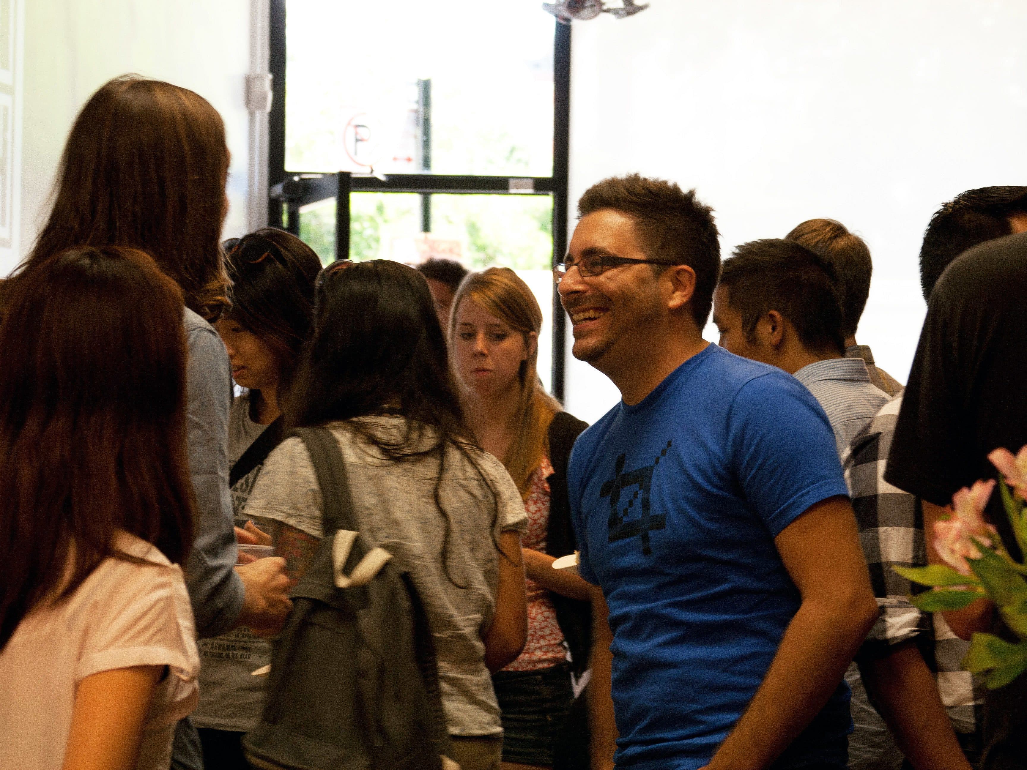 A group of people interacting at an indoor social gathering. A man in a blue shirt with glasses is smiling and talking to others. Several individuals are engaged in conversation, and a few are facing away from the camera. Flowers are visible in the foreground.