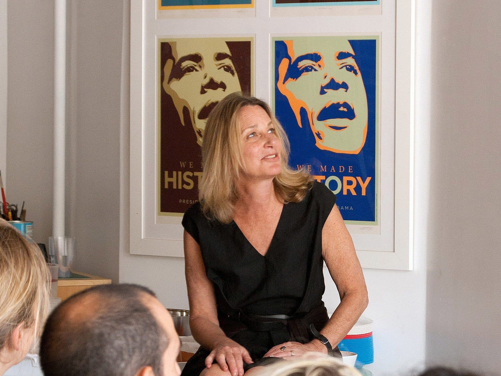 A woman with blonde hair, wearing a black dress, is speaking to a small group of people. Behind her are framed posters with stylized images of former U.S. President Barack Obama. The setting appears to be indoors, possibly in a discussion or presentation setting.