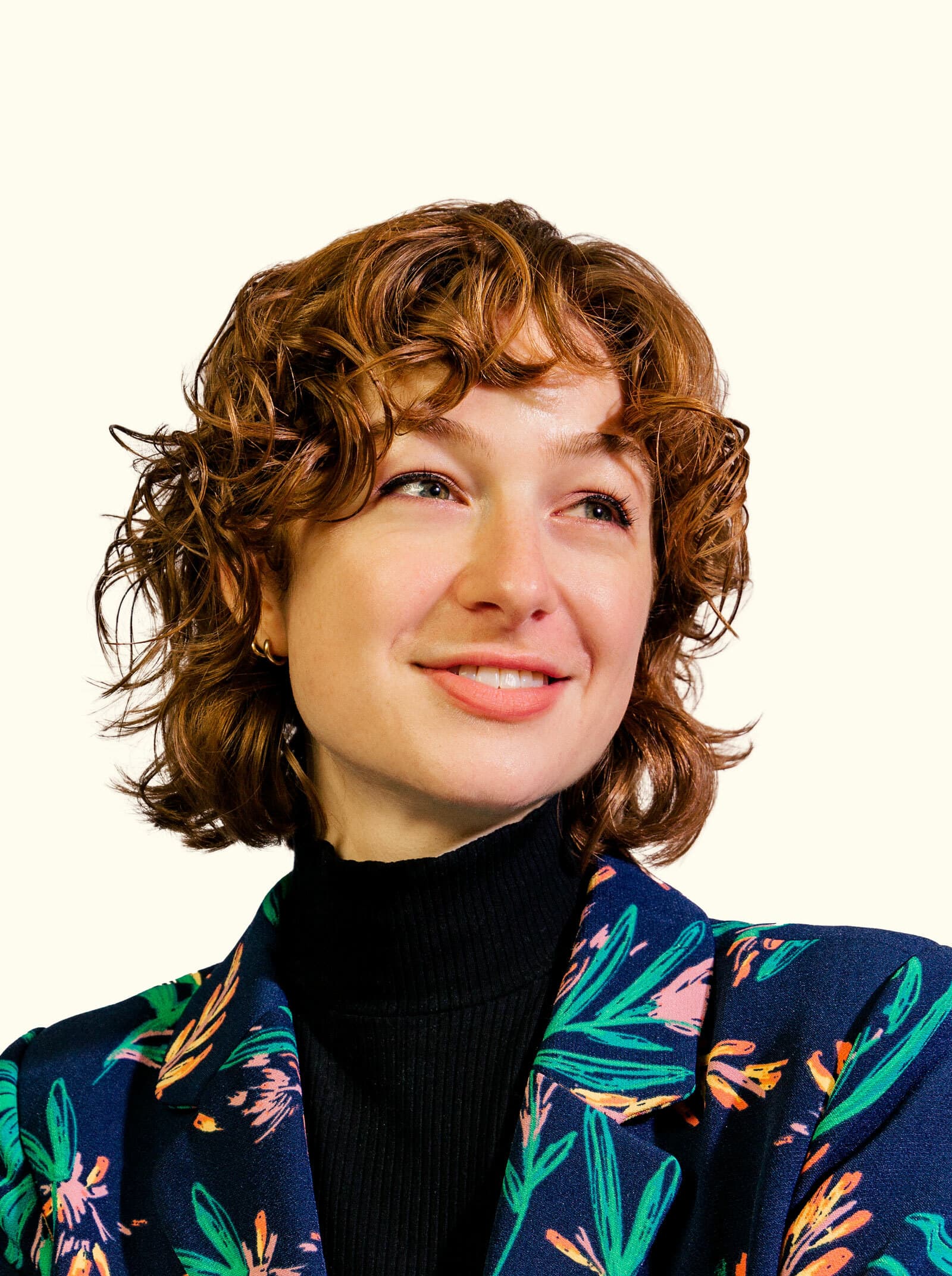 A person with curly auburn hair smiles while looking slightly to the side. They are wearing a black turtleneck and a dark, floral-patterned blazer, set against a plain light background.